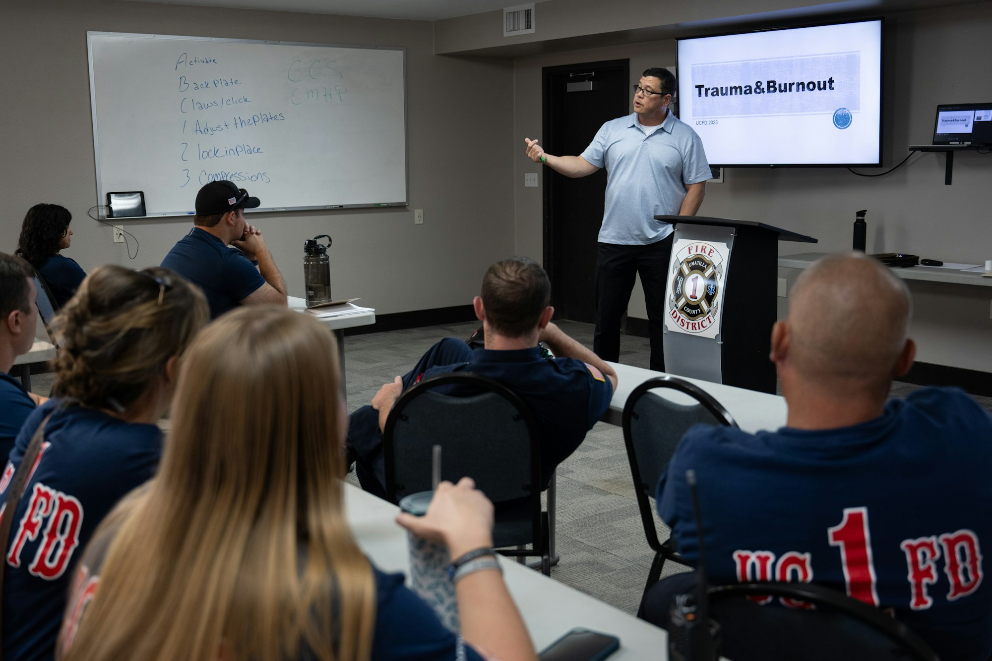 A speaker addresses firefighters about "Trauma & Burnout" in a classroom setting with a whiteboard and screen.