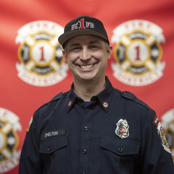 A smiling firefighter in uniform with badges and a cap, in front of a backdrop with fire department emblems.