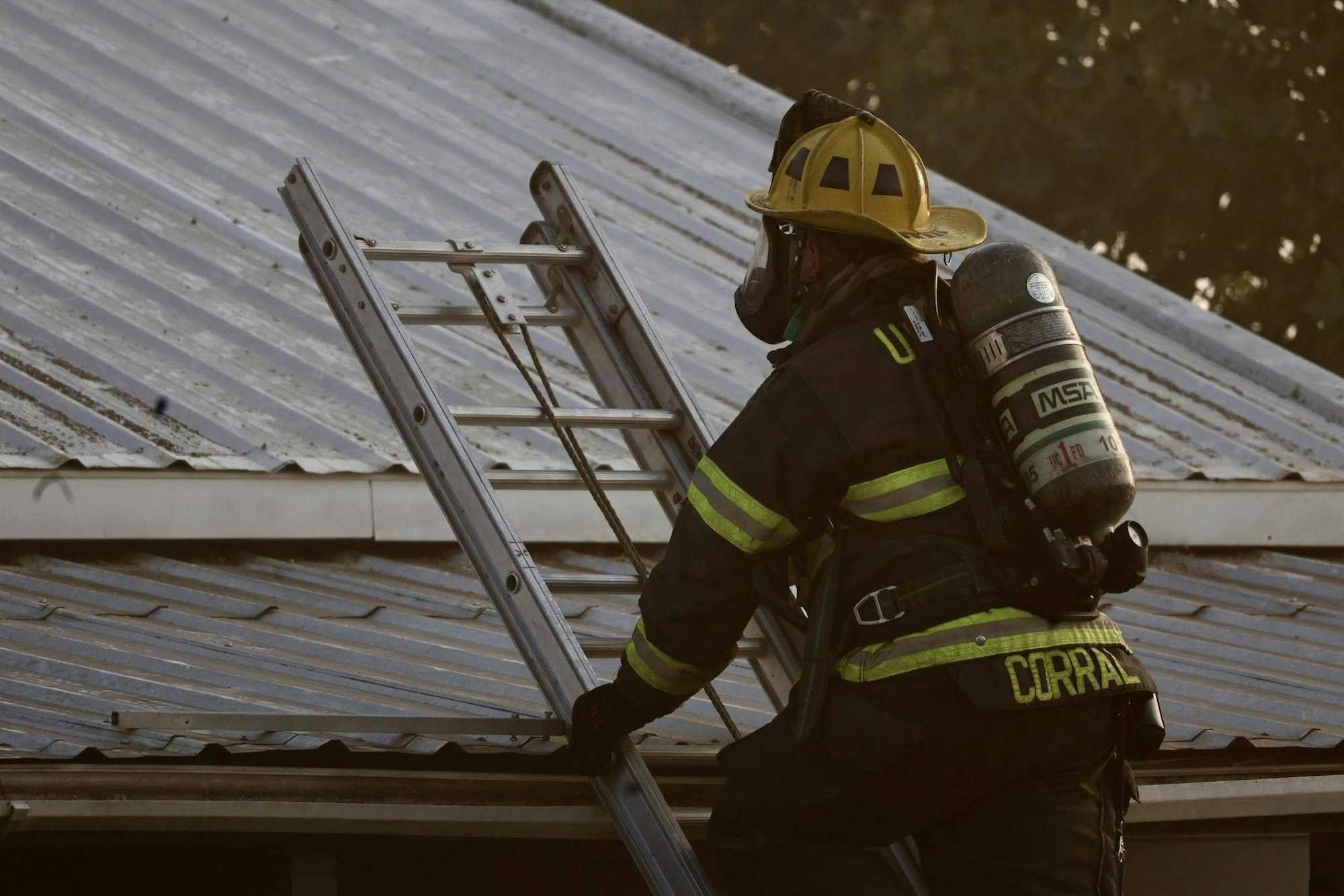 Firefighter in gear climbing a ladder onto a metal roof.
