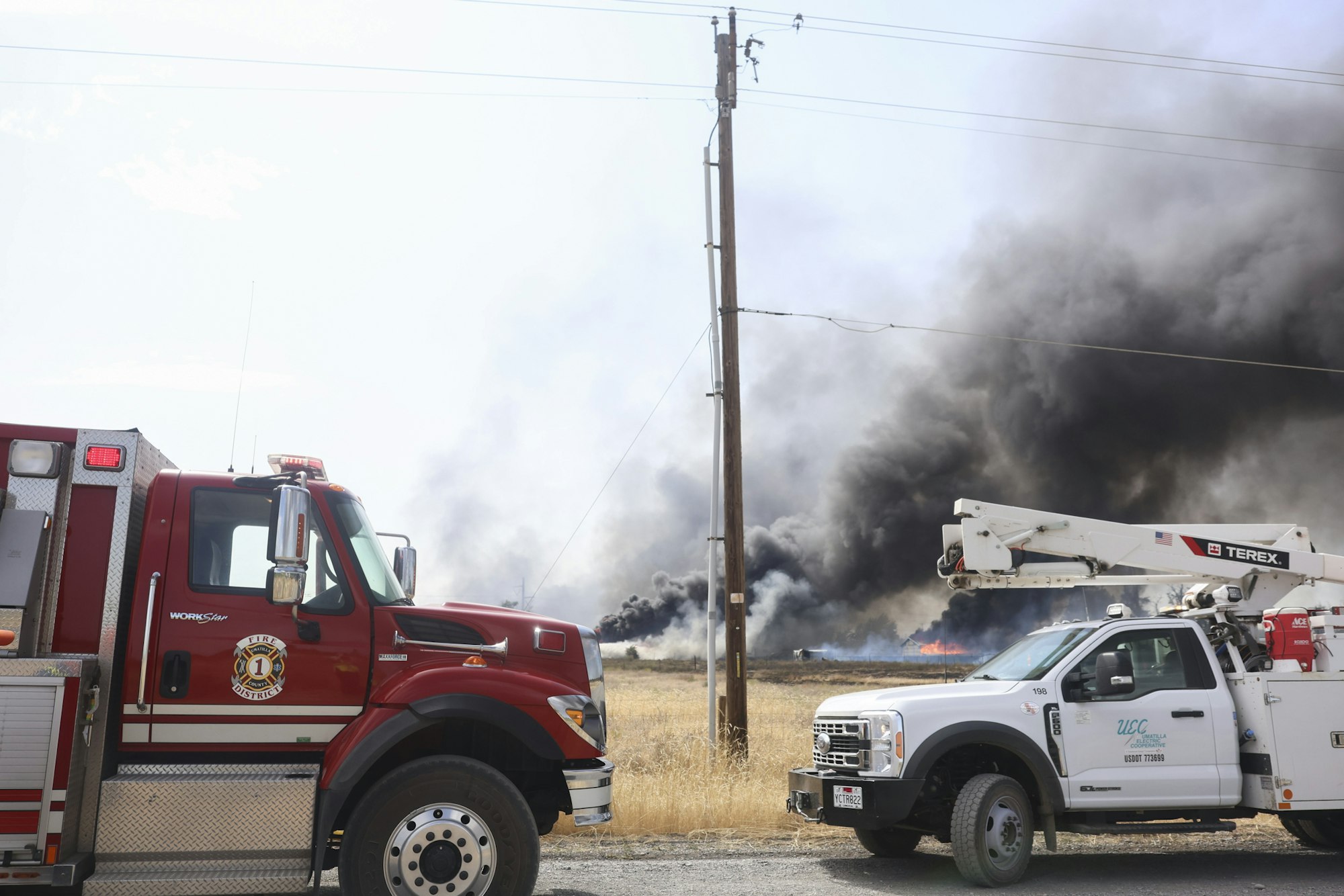 Fire truck and utility truck near smoke and flames in a field fire.