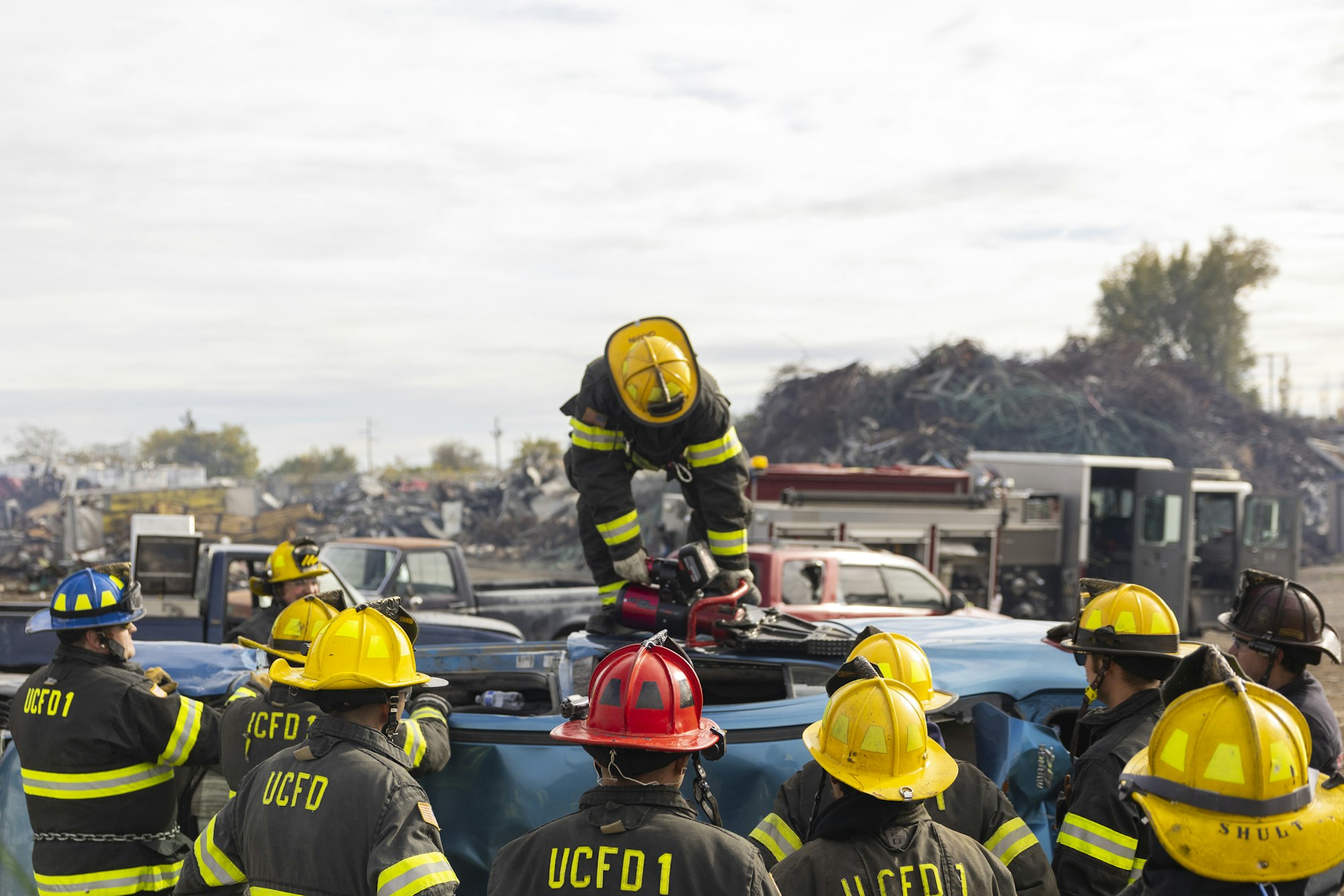 Firefighters in gear are practicing a rescue drill, using tools on a flipped vehicle.