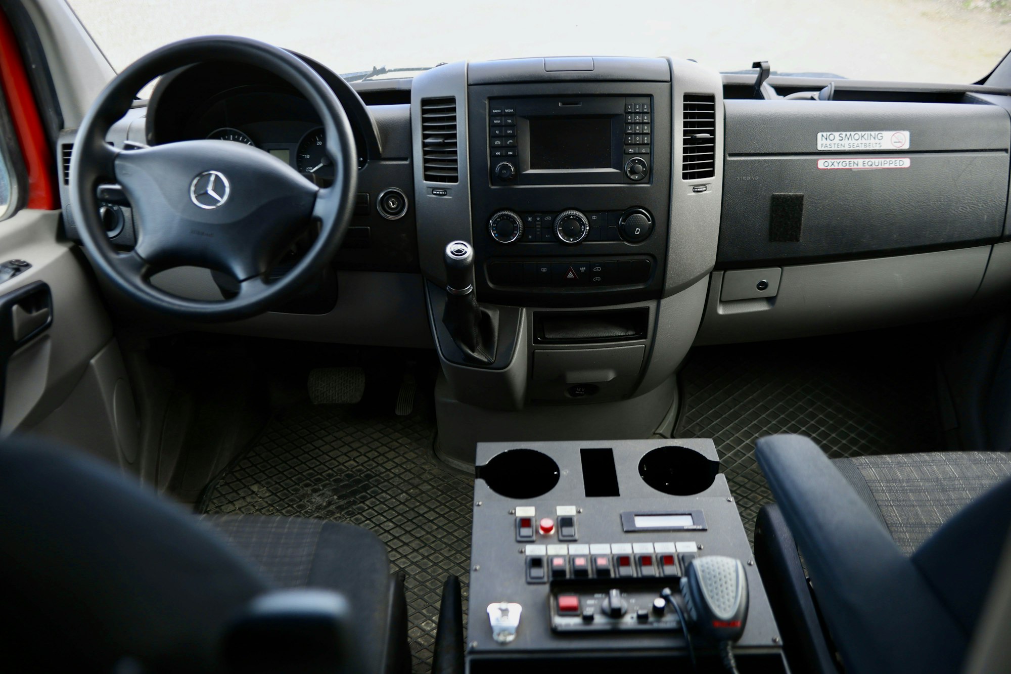 Dashboard and steering wheel of a Mercedes vehicle, with control panel and radio/mic. "No Smoking" sign.