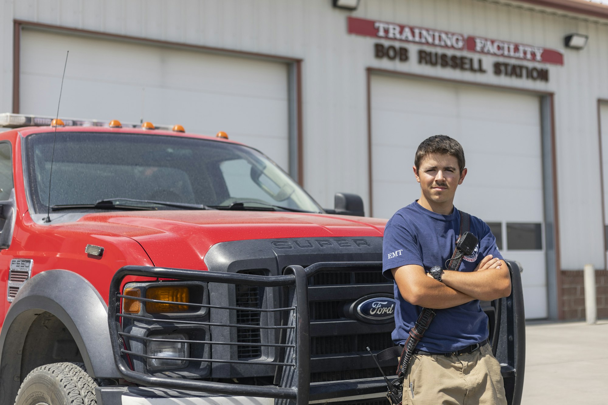 A person in an EMT shirt stands by a red Ford F-550 truck outside a training facility labeled "Bob Russell Station."