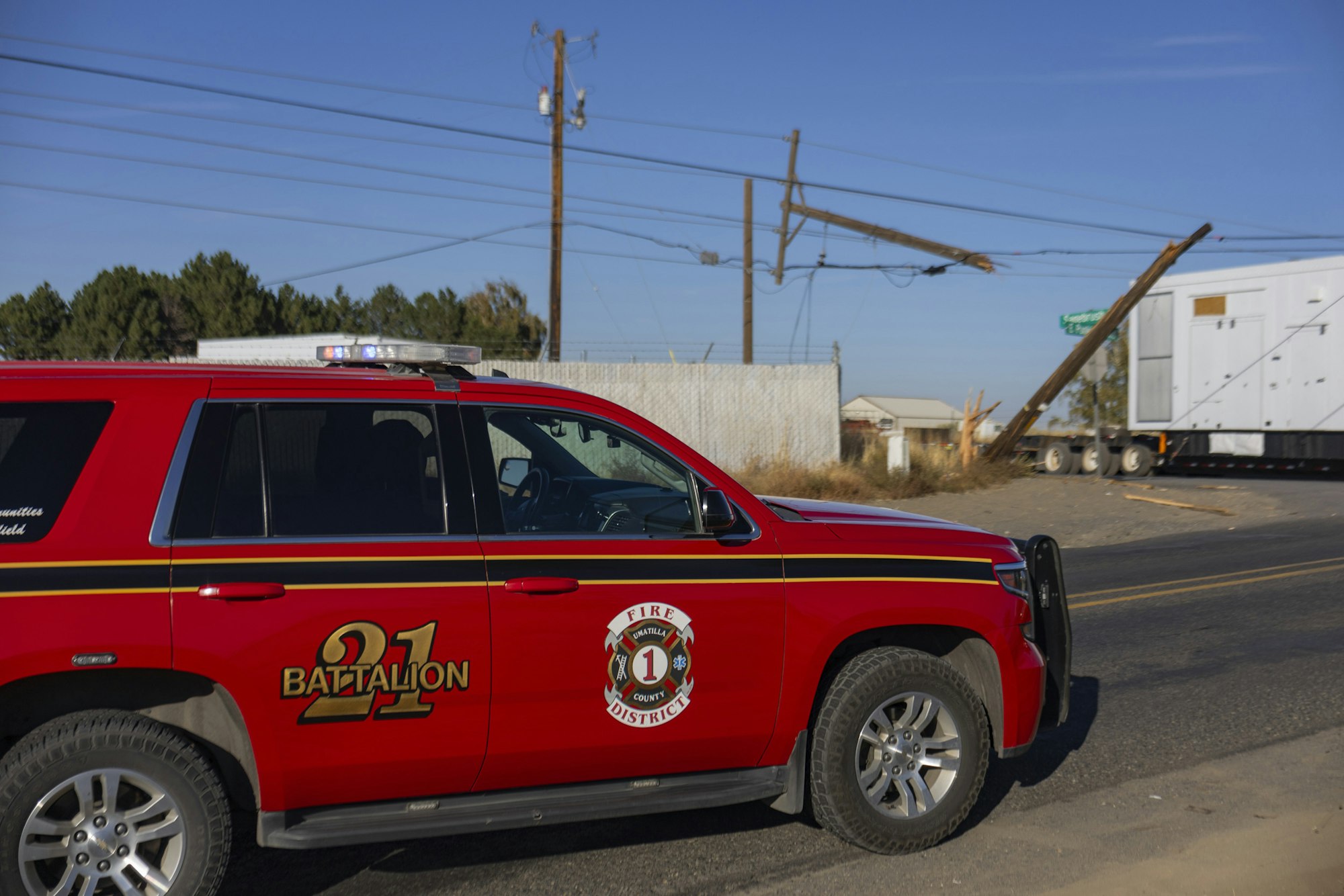 A red battalion fire vehicle near a broken power pole and a large trailer on the road.