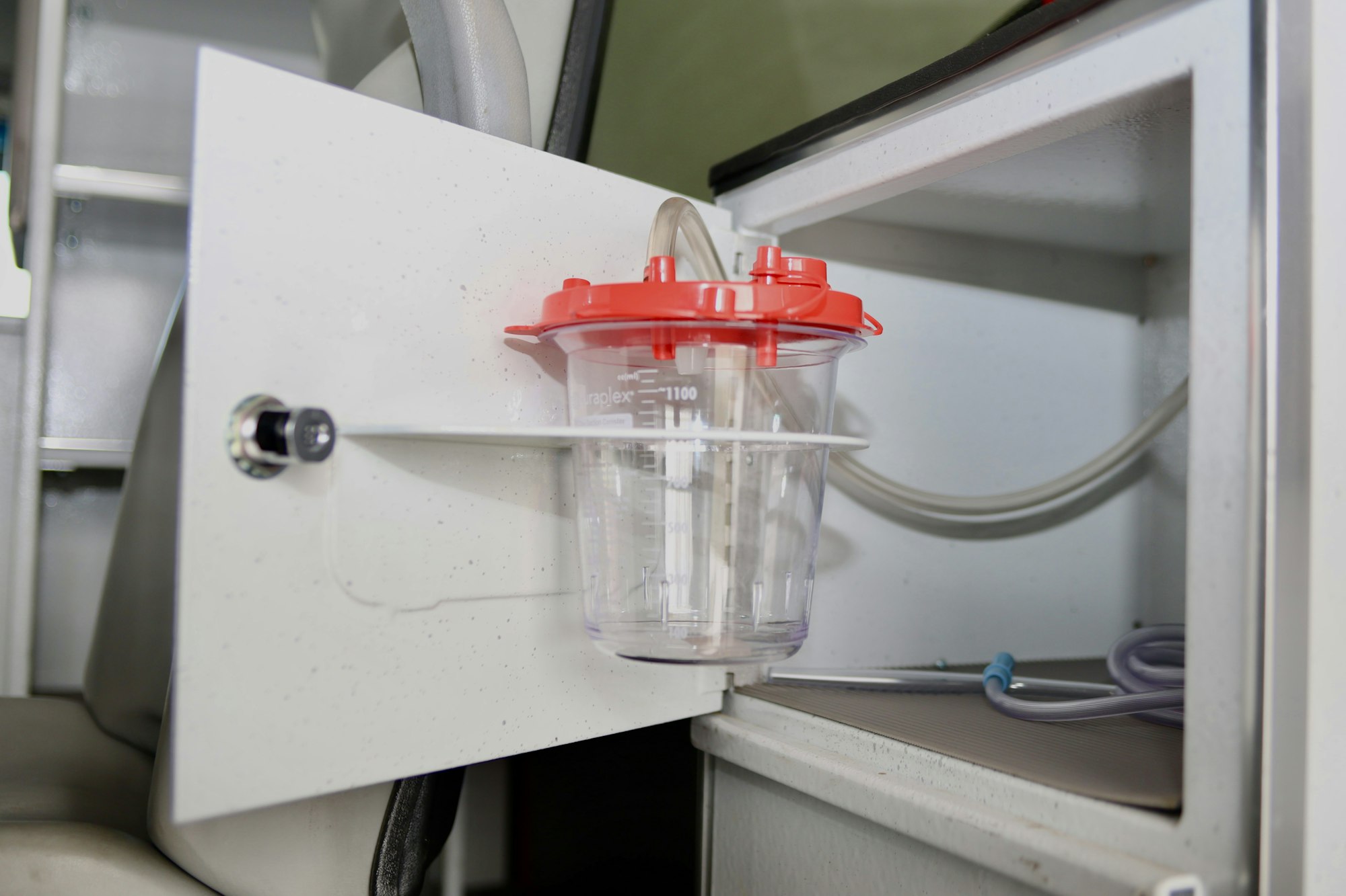 A medical suction canister with a red lid is attached inside a cabinet, connected to tubes.