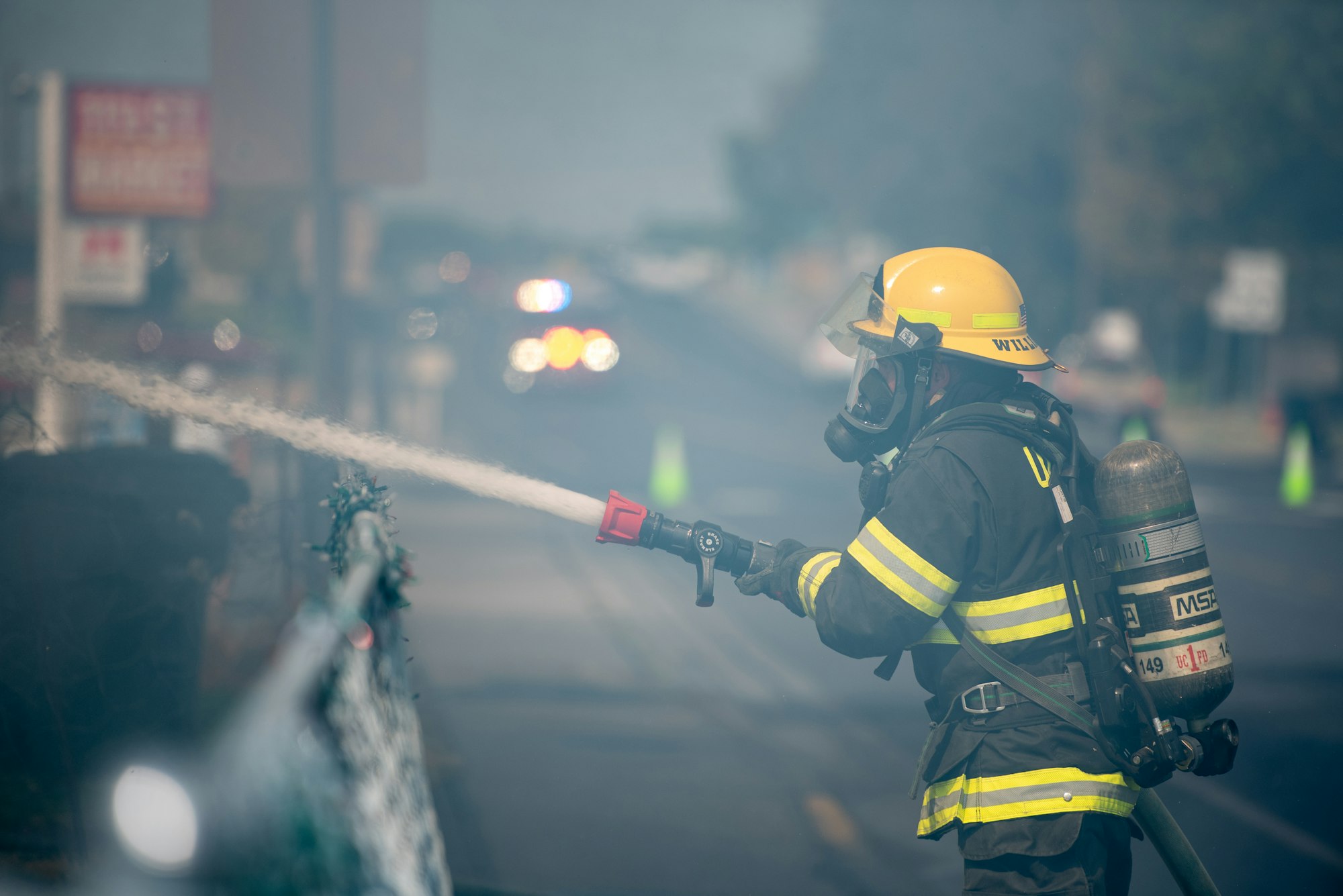 Firefighter in gear sprays water from a hose onto a smoky area near a fence, blurred emergency lights in the background.