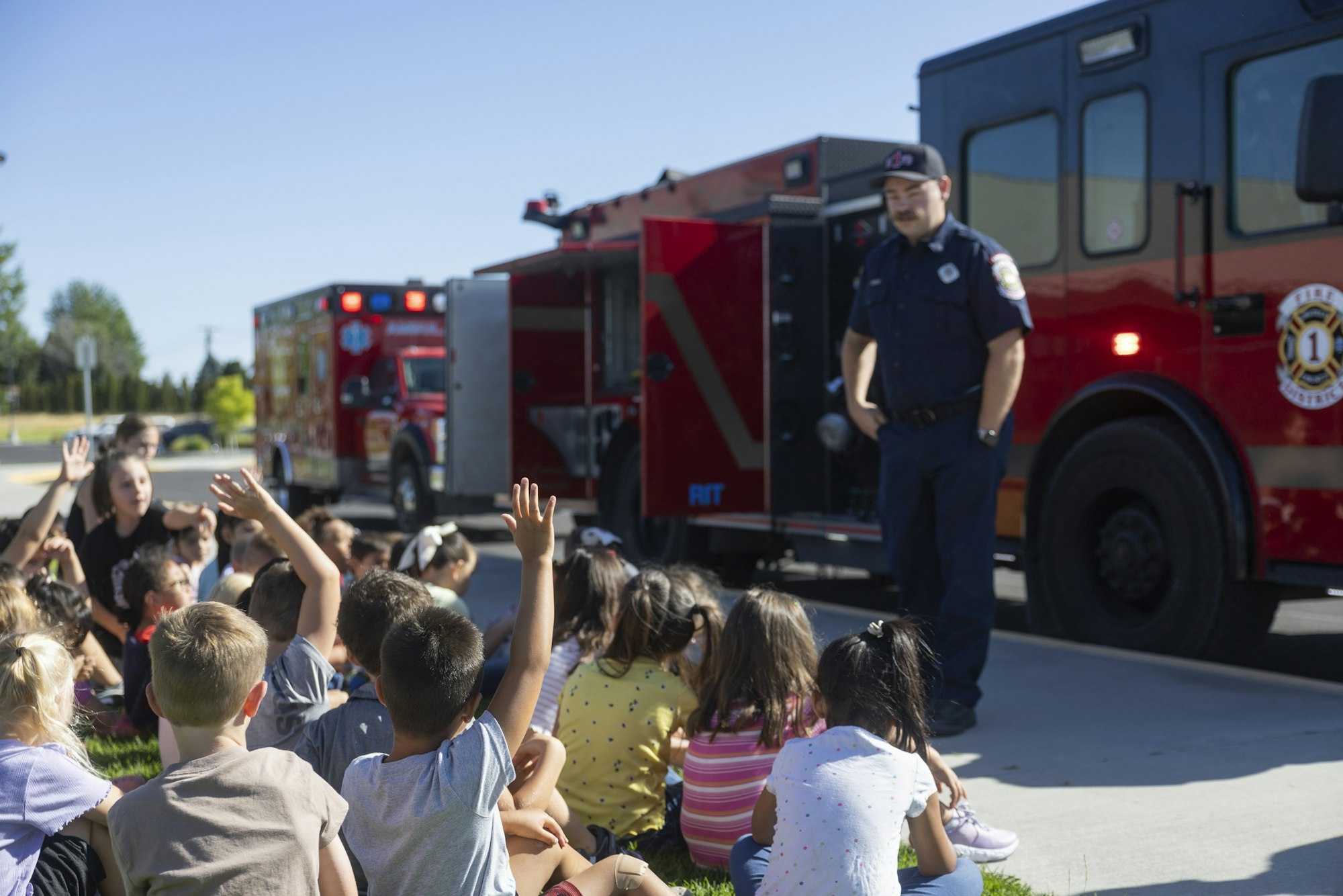 A firefighter talks to a group of children sitting on the ground near fire trucks.