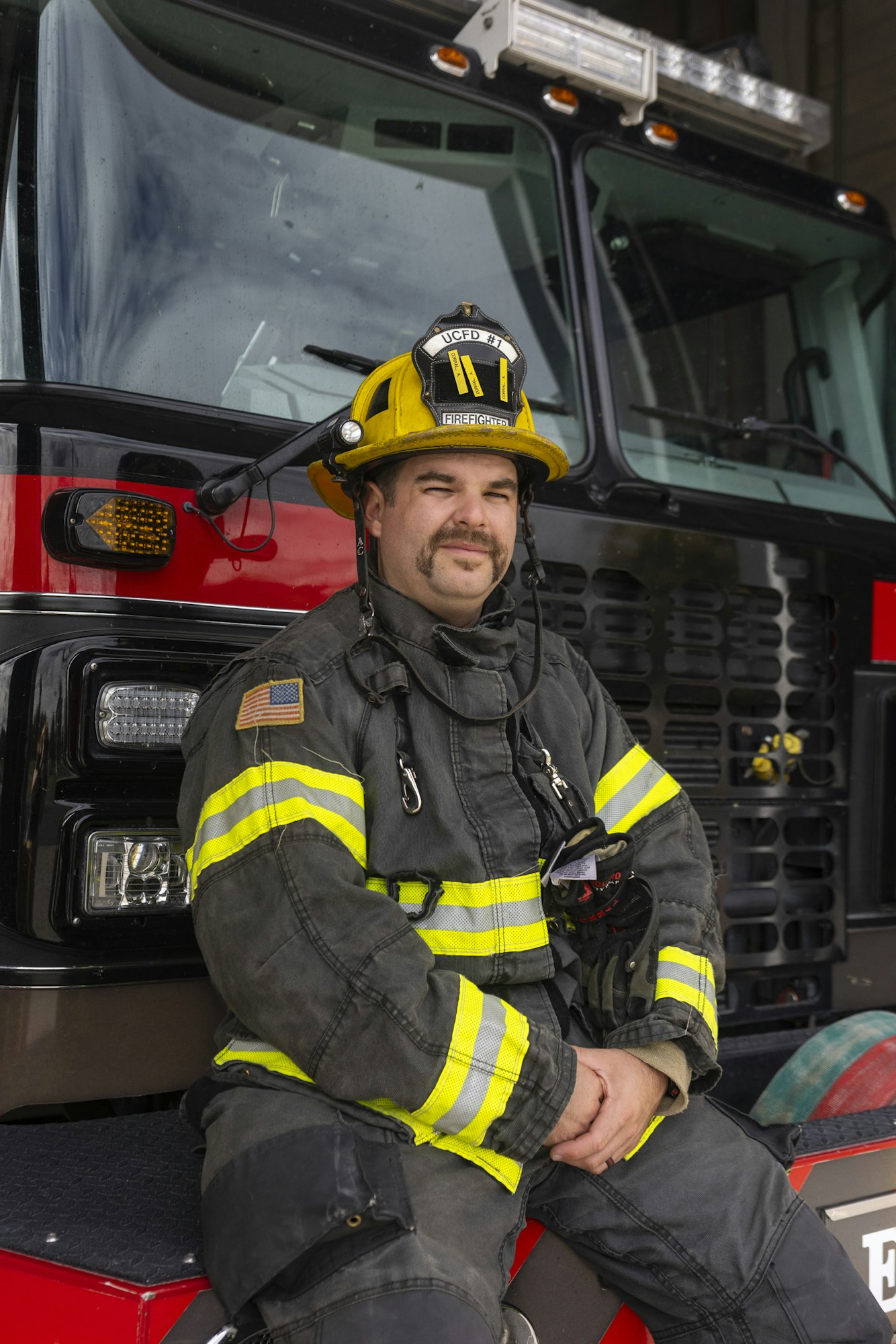 A firefighter in uniform and helmet sits on a fire truck, looking at the camera.