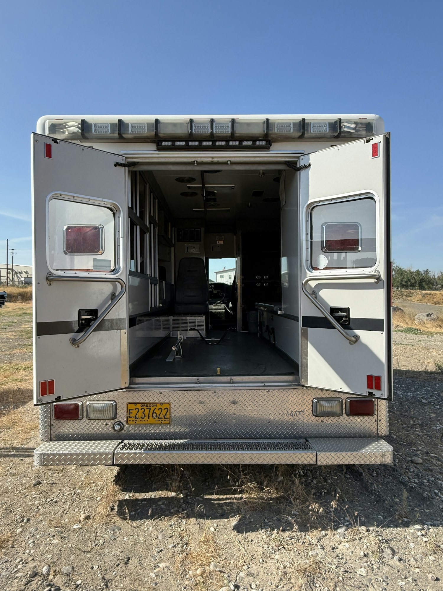 The image shows the open rear of an empty ambulance on a gravel area with a clear blue sky.