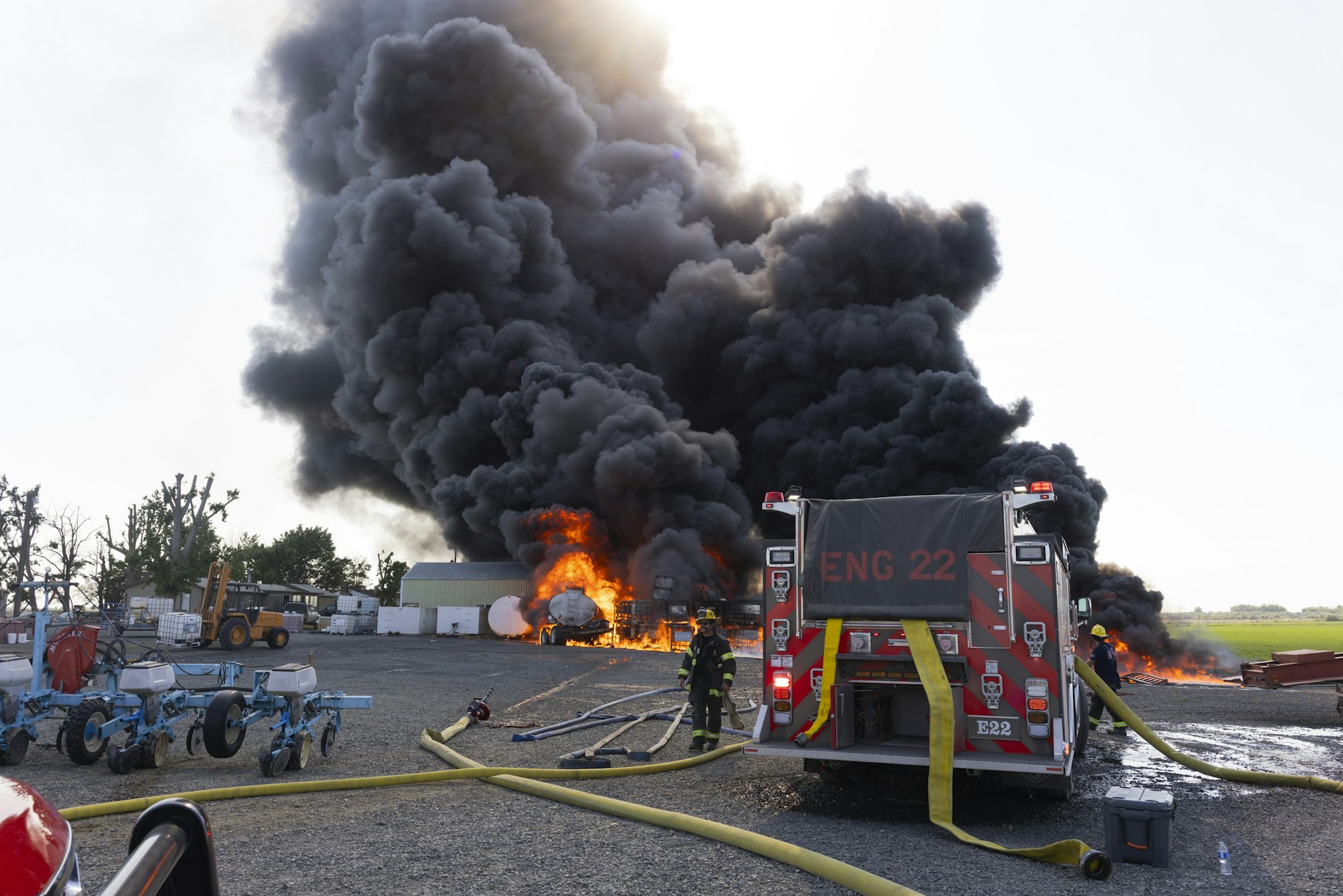 Firefighters battle a large fire with heavy smoke at a site, using fire hoses near a fire engine.