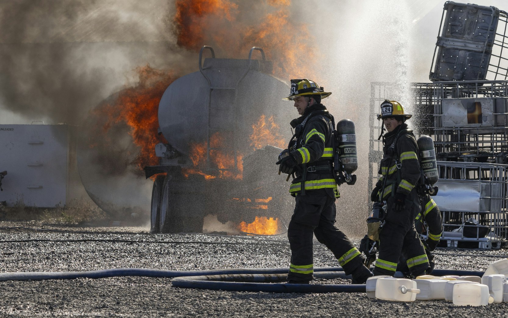Firefighters in gear walk past a burning truck with thick smoke and flames, surrounded by equipment.