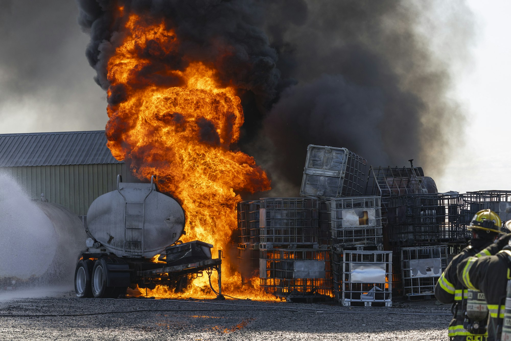 Firefighters tackling a massive blaze involving a burning tanker and storage containers, with thick smoke engulfing the scene.