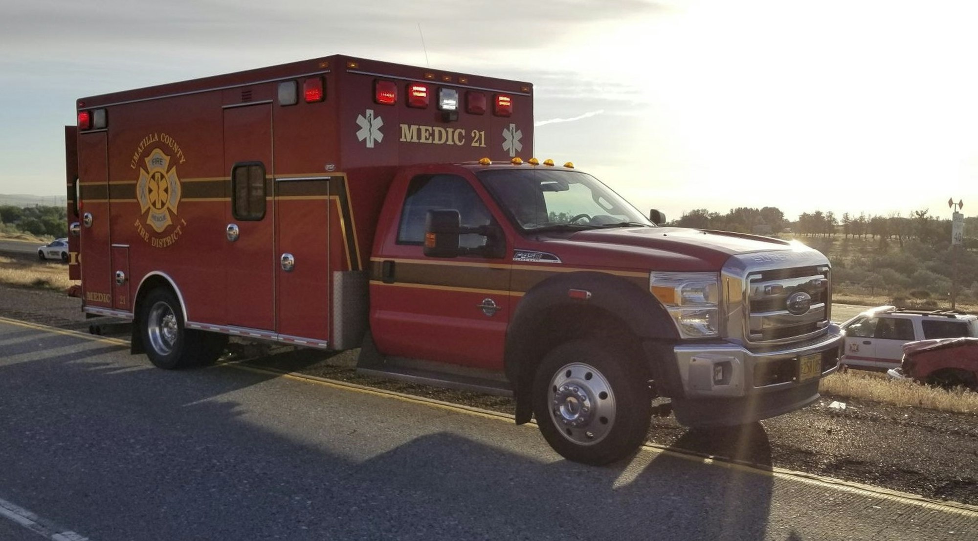 A red ambulance parked on a roadside during sunset, marked "MEDIC 21," with scenic background and emergency lights.