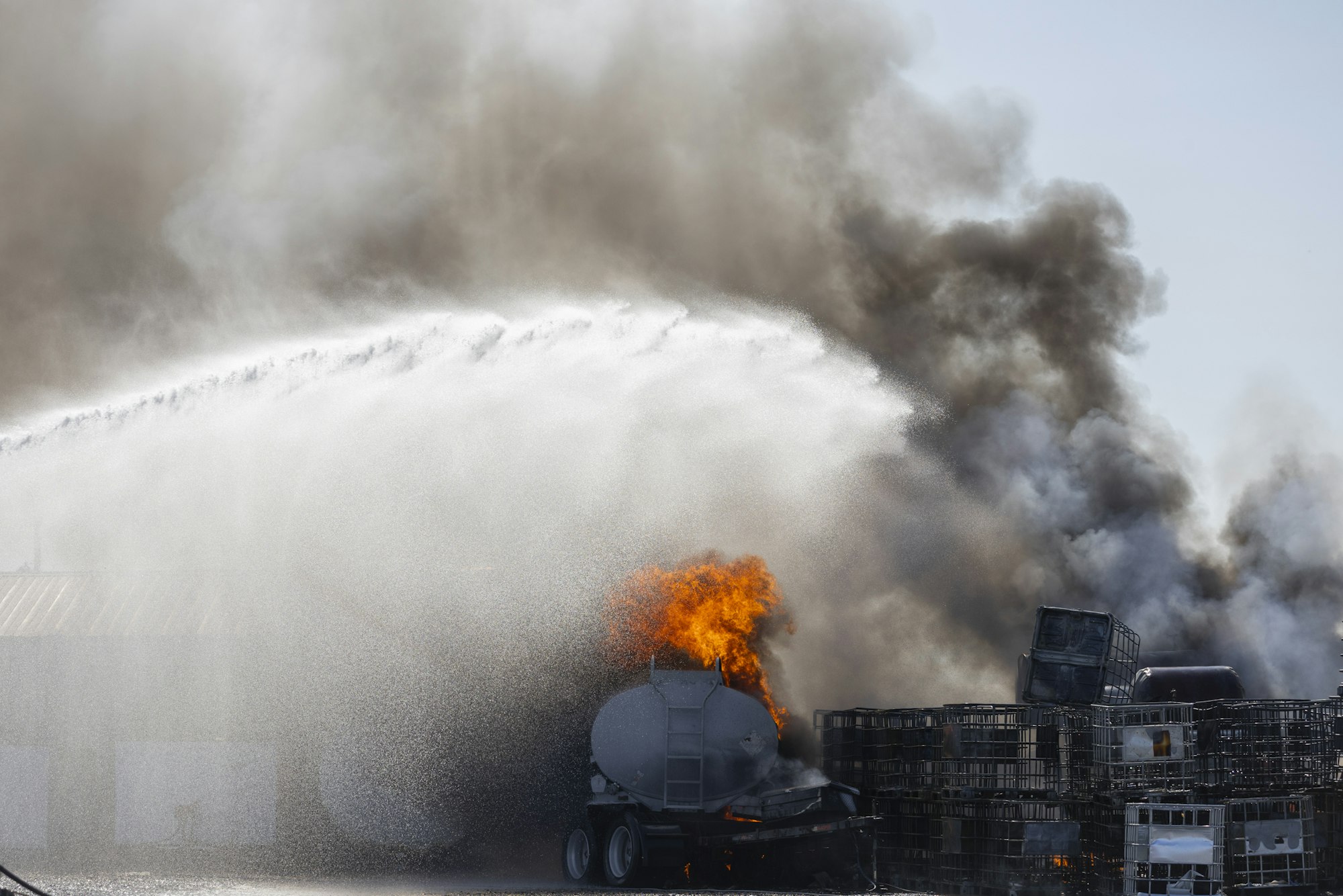 A fire being doused with water, smoke billows, and orange flames are visible near containers.