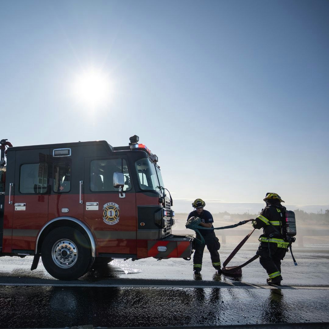 Firefighters working with a hose near a fire truck under the sun.