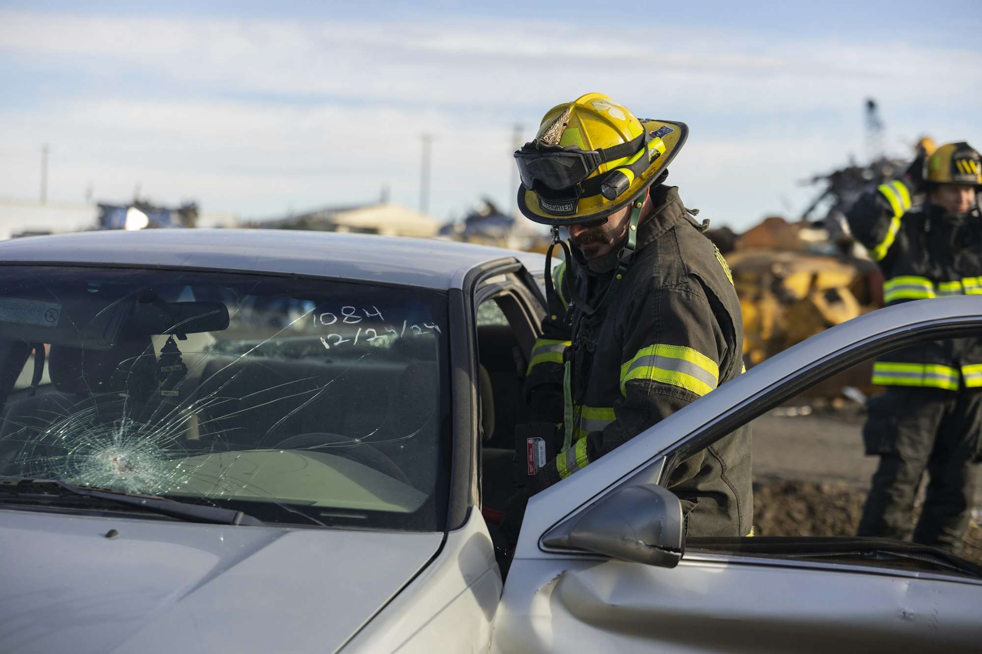 Firefighter inspecting a car with a cracked windshield, wearing yellow helmet and protective gear.