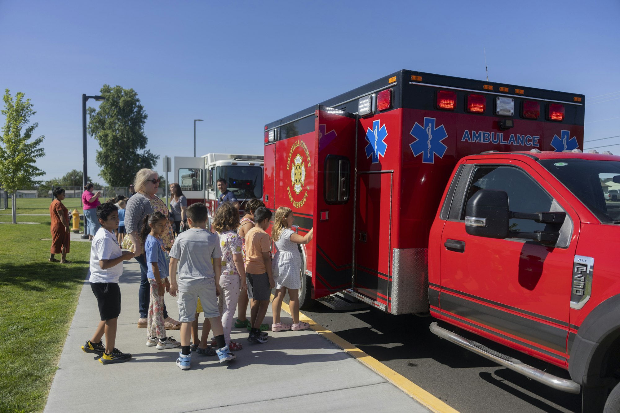 A group of children and adults gather around a red ambulance parked on a sunny day.