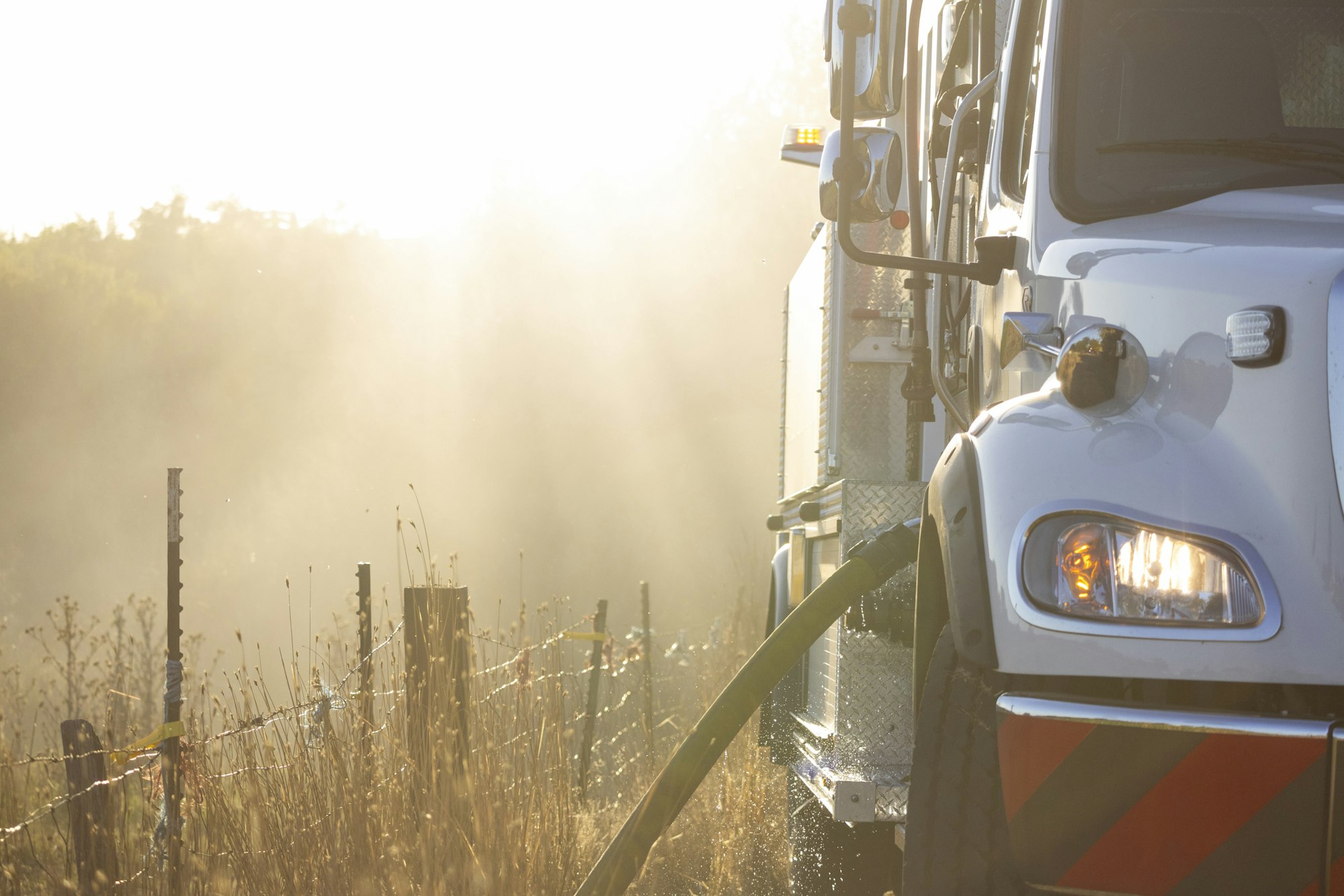 A truck with a hose in a rural area, surrounded by sunlight and dust near a wire fence.