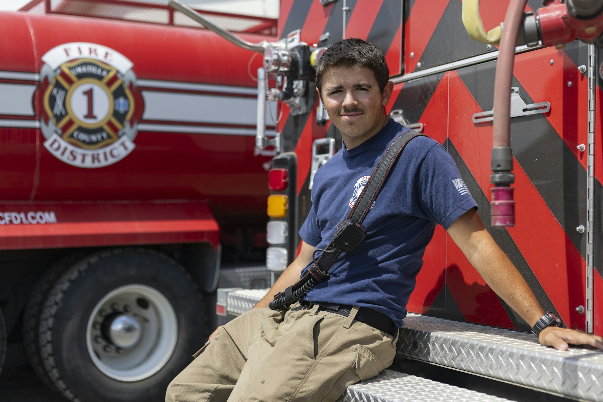 A firefighter sitting on a fire truck, wearing a navy shirt and khaki pants, with a portable radio.