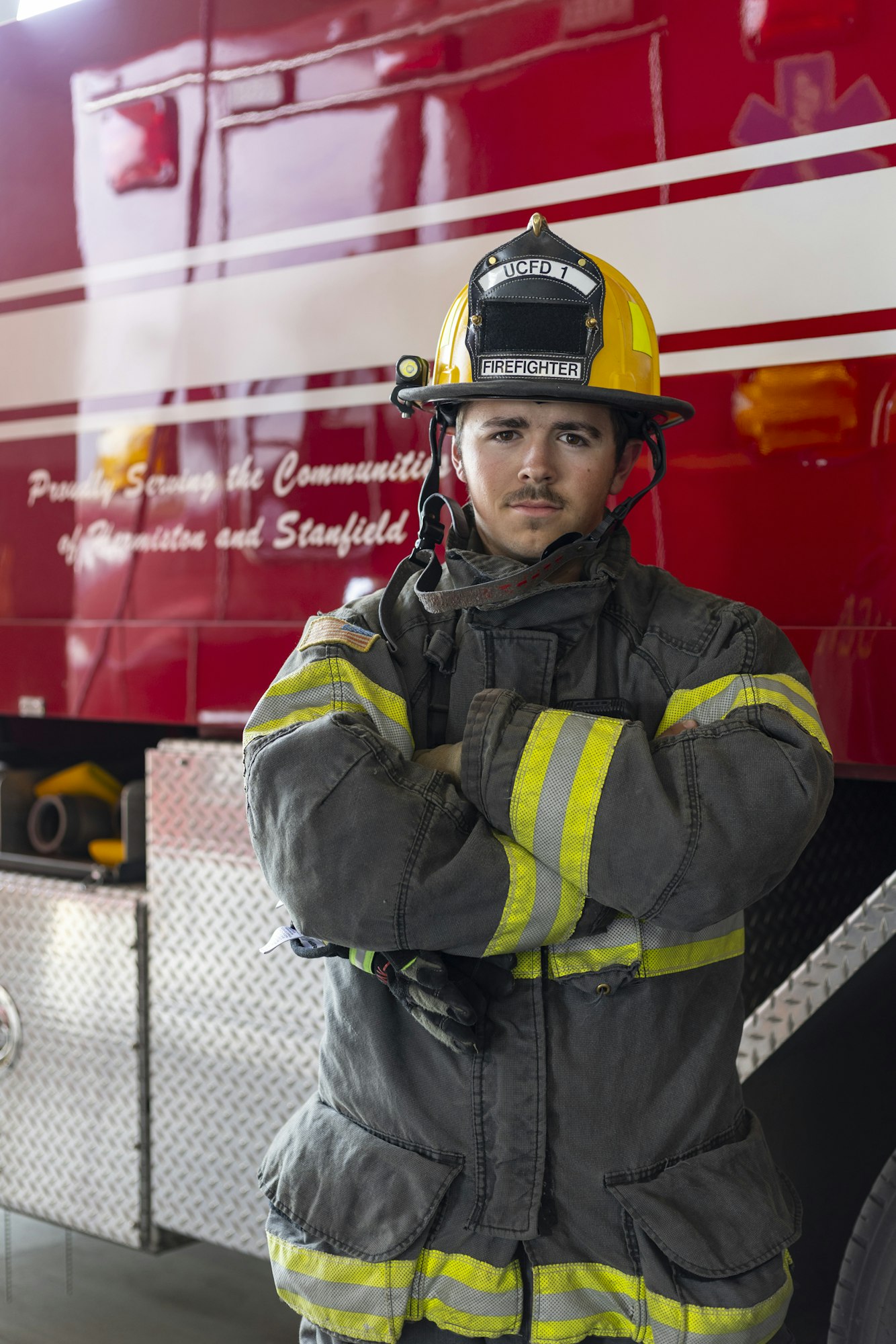 A firefighter in uniform stands in front of a red fire truck, arms crossed, with UCFD 1 on helmet.