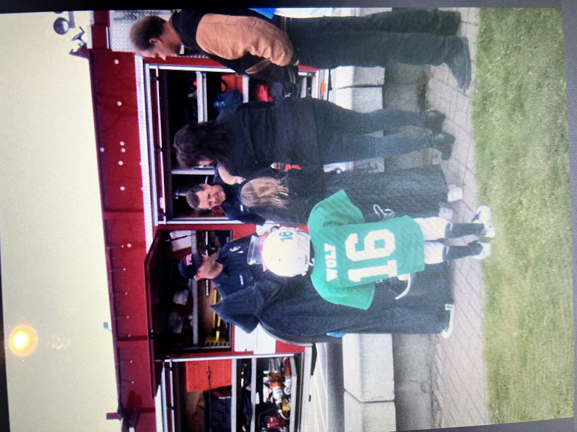 A child in sports gear, wearing a green jersey with number 16, stands near people by a fire truck.