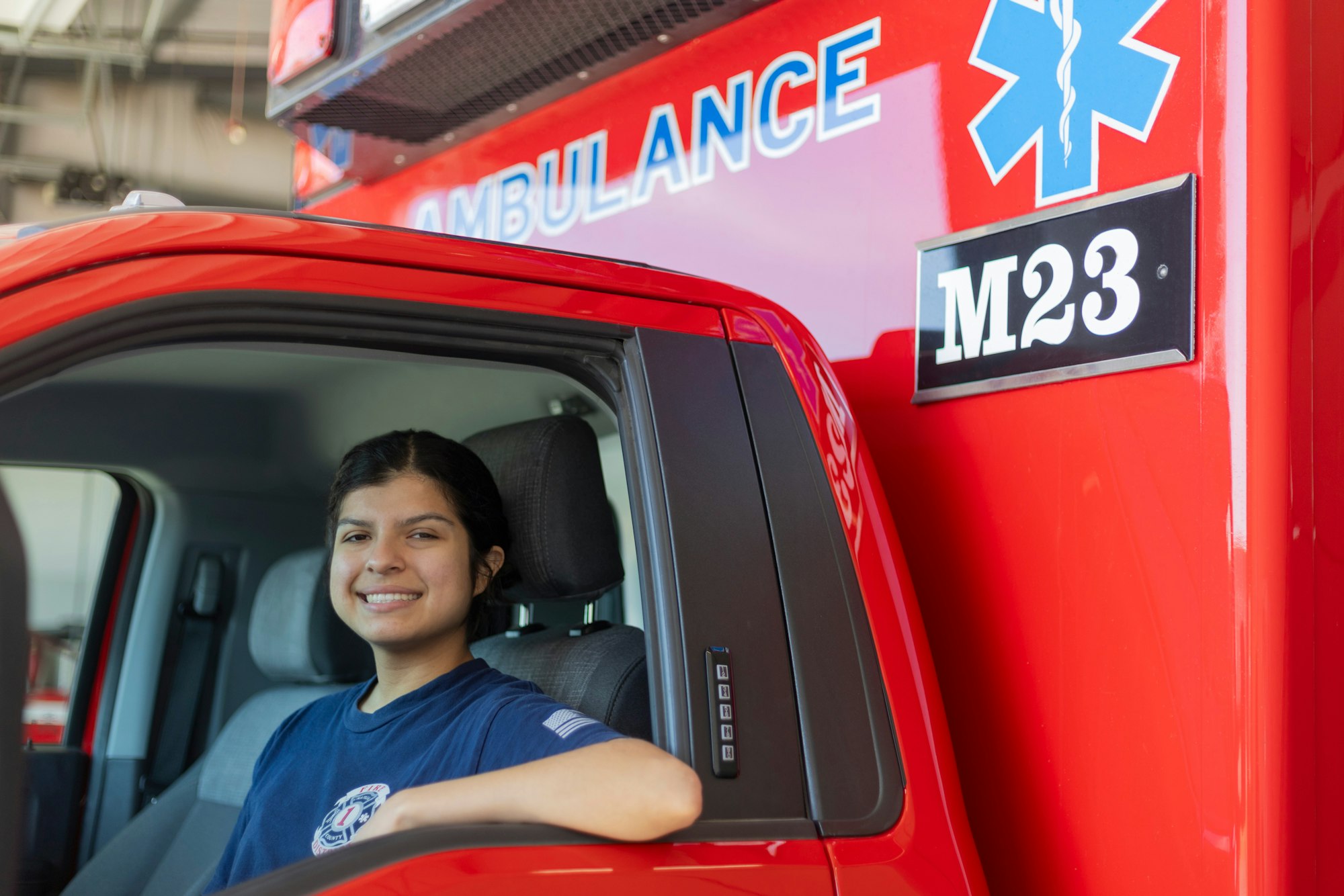 Person smiling inside a red ambulance with "AMBULANCE" and "M23" visible on the side.