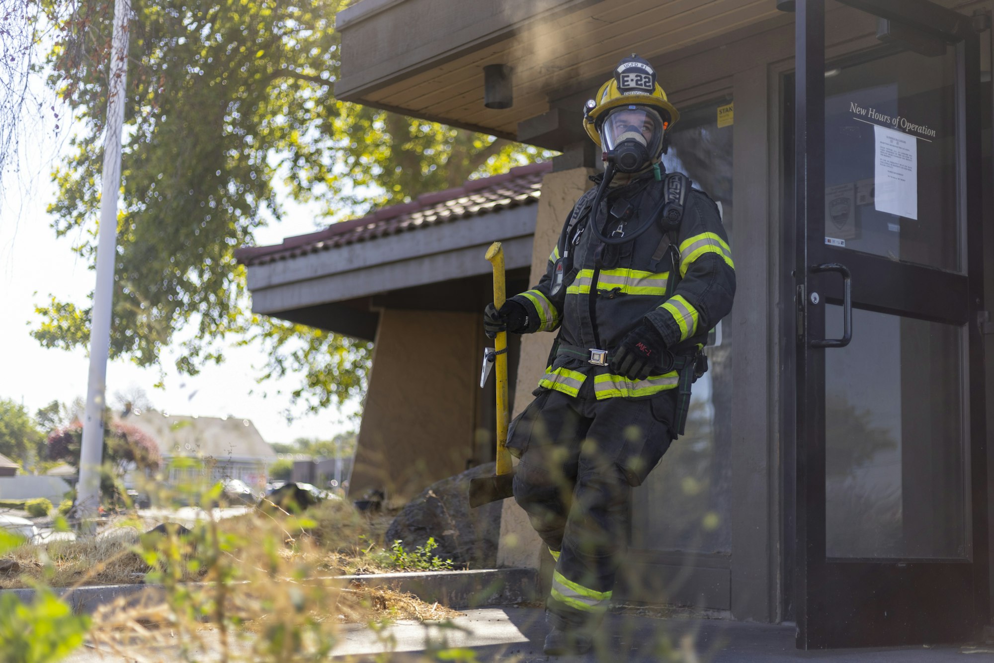 A firefighter in full gear, holding an axe, exits a building on a sunny day.