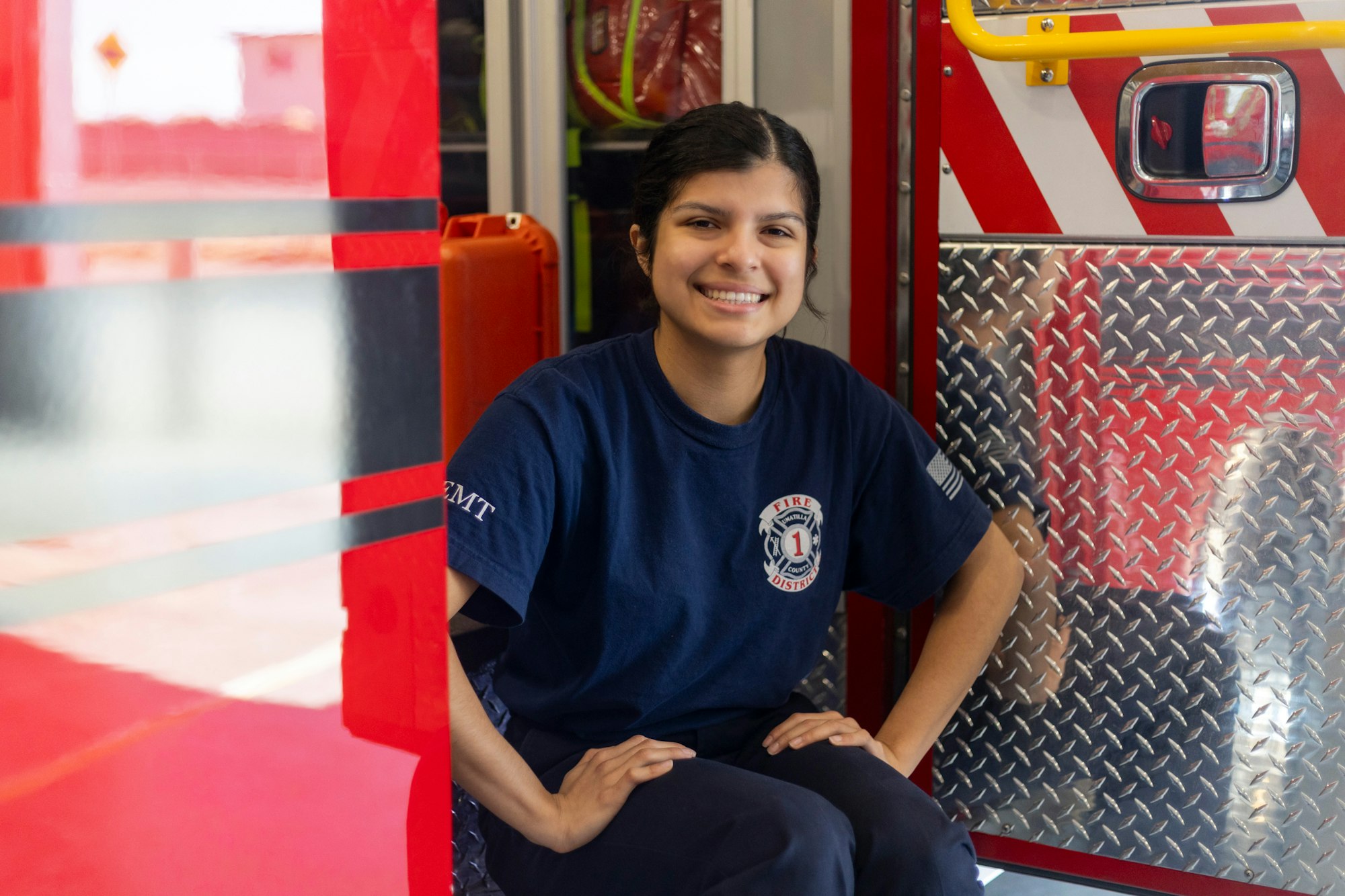 A person in an EMT shirt smiling, sitting inside a fire truck with red and silver details.
