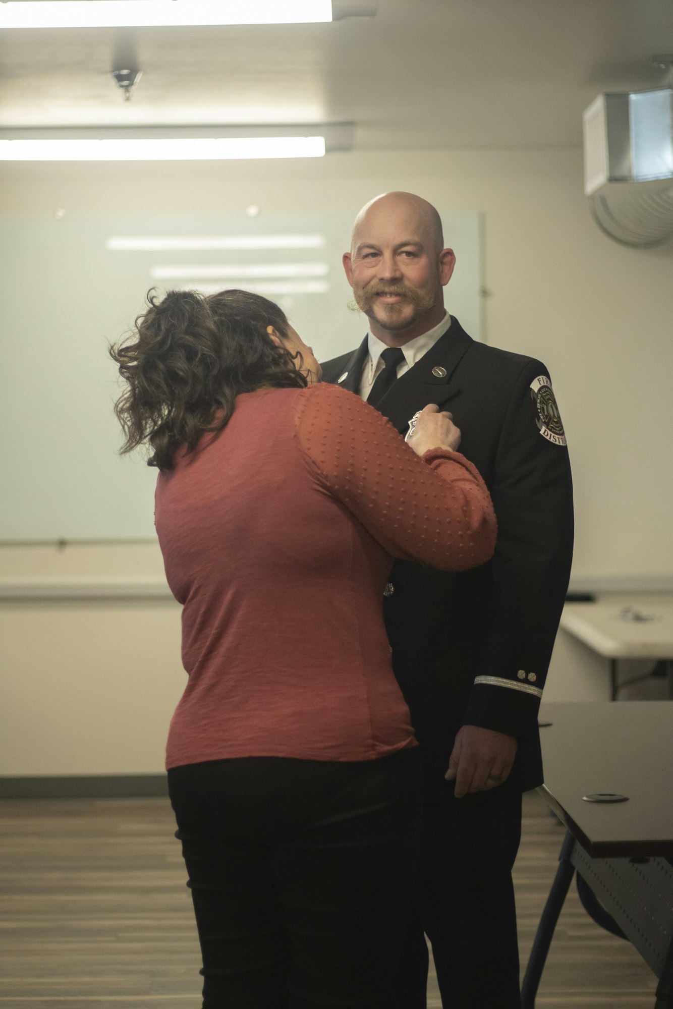 A person in a red shirt pins a badge on a uniformed individual in a room.