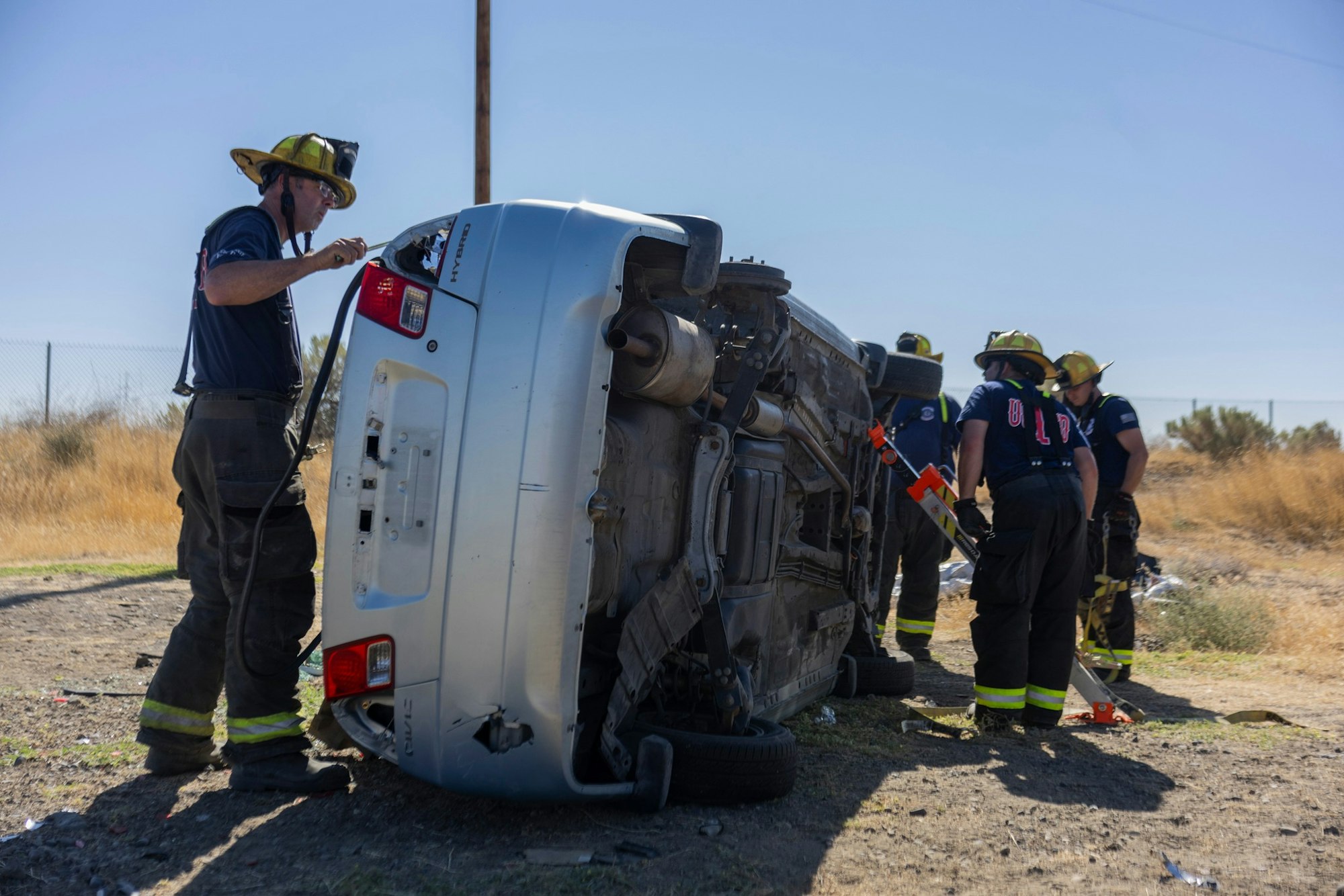Firefighters assess a car overturned on its side in a dry grass area.