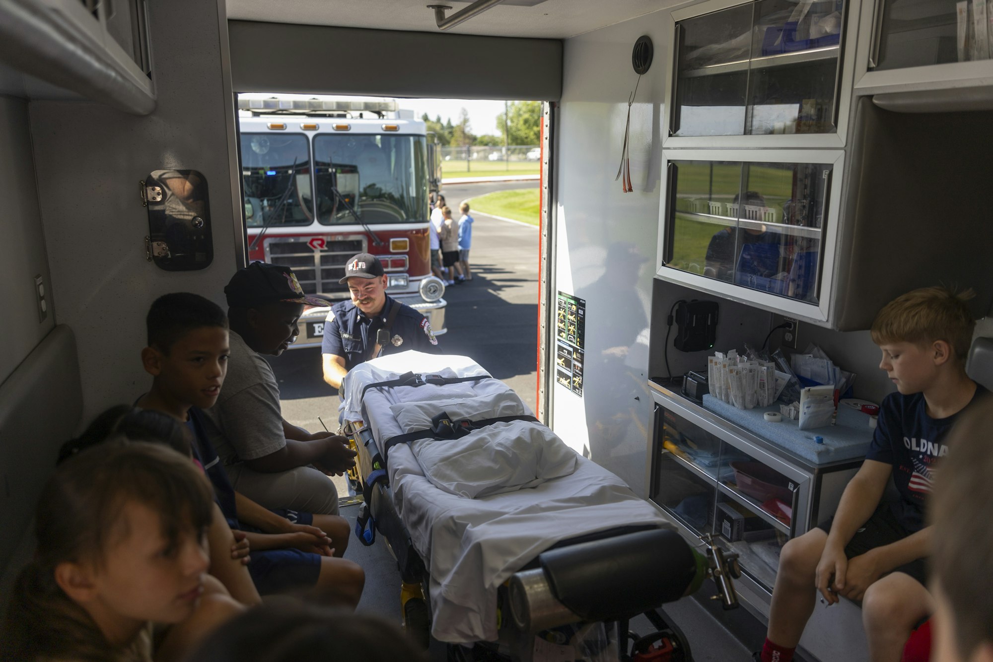 Children sitting inside an ambulance with a firefighter nearby, fire truck visible outside.