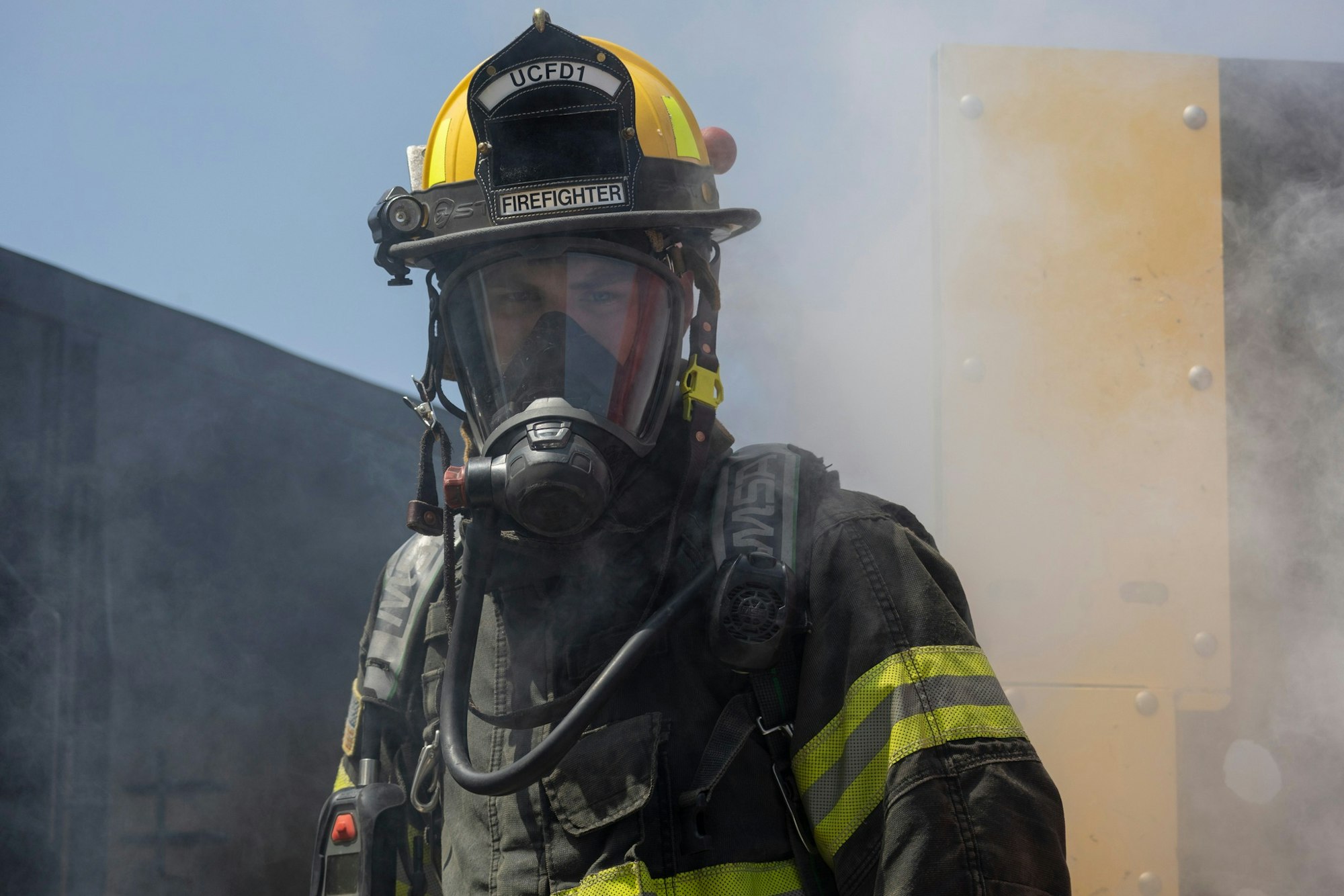 A firefighter in full gear and breathing apparatus stands amidst smoke.