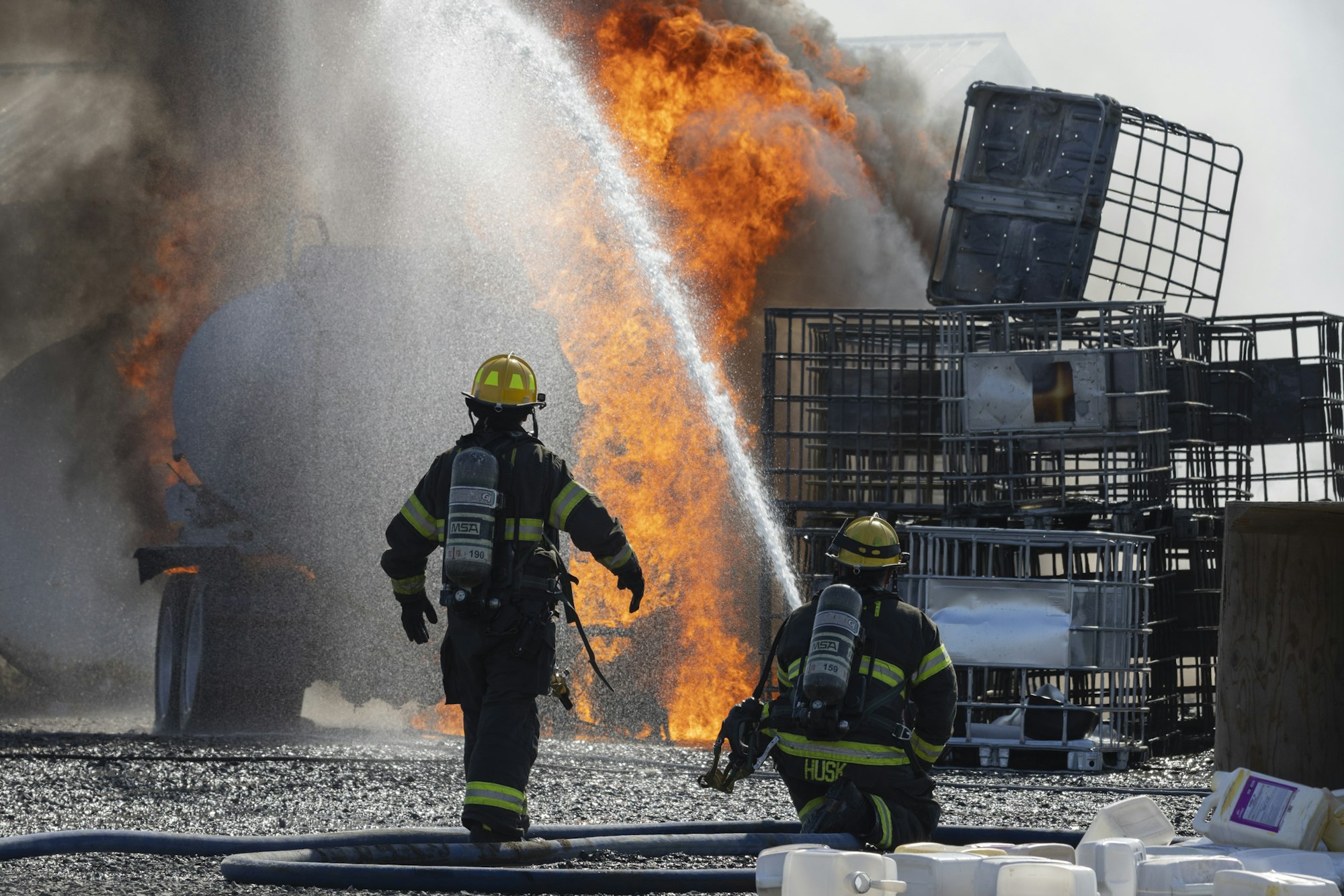 Firefighters in protective gear spray water on a large outdoor fire with stacked metal cages nearby.