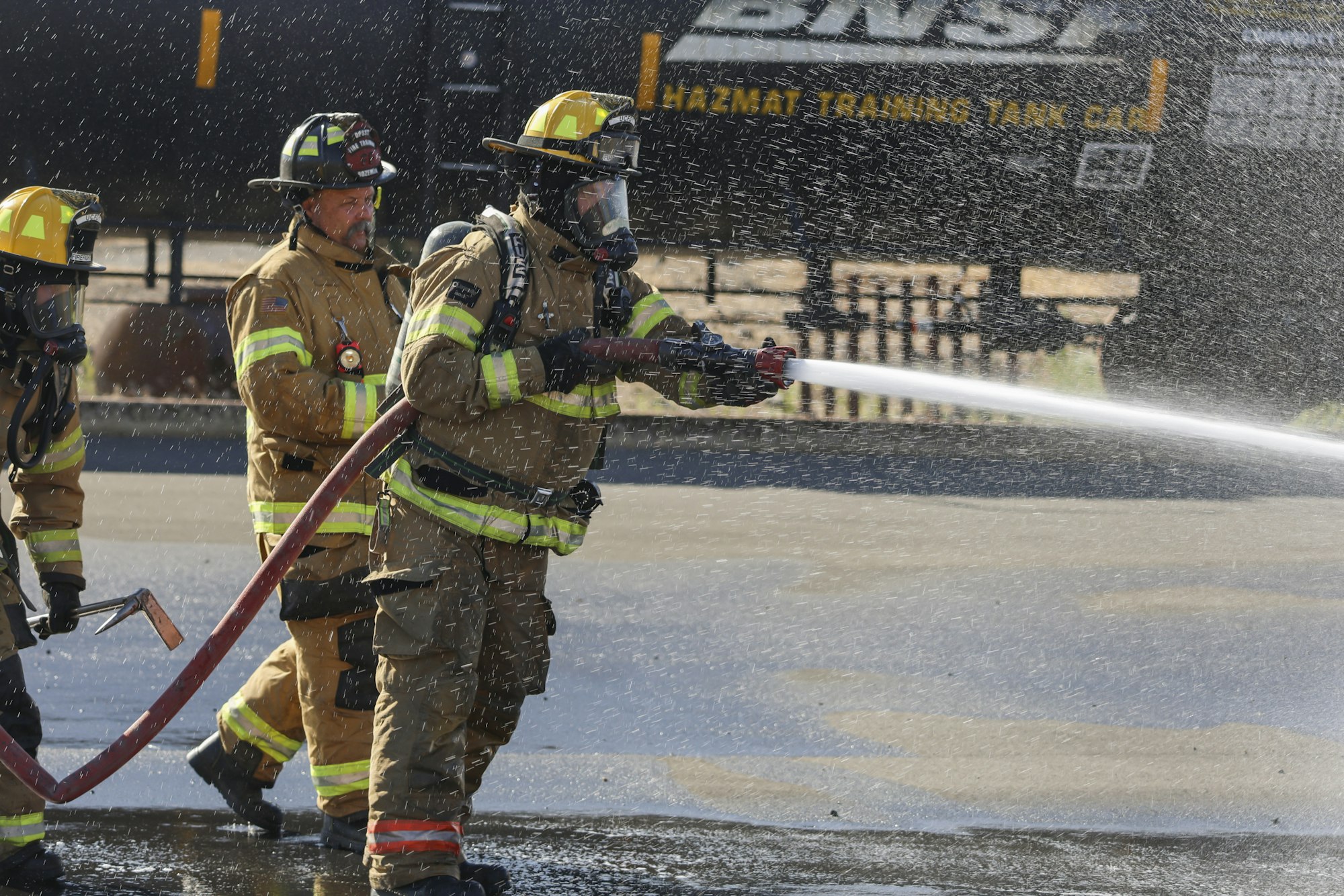 Firefighters in gear spray water from a hose during a training exercise with a hazmat tank car in the background.