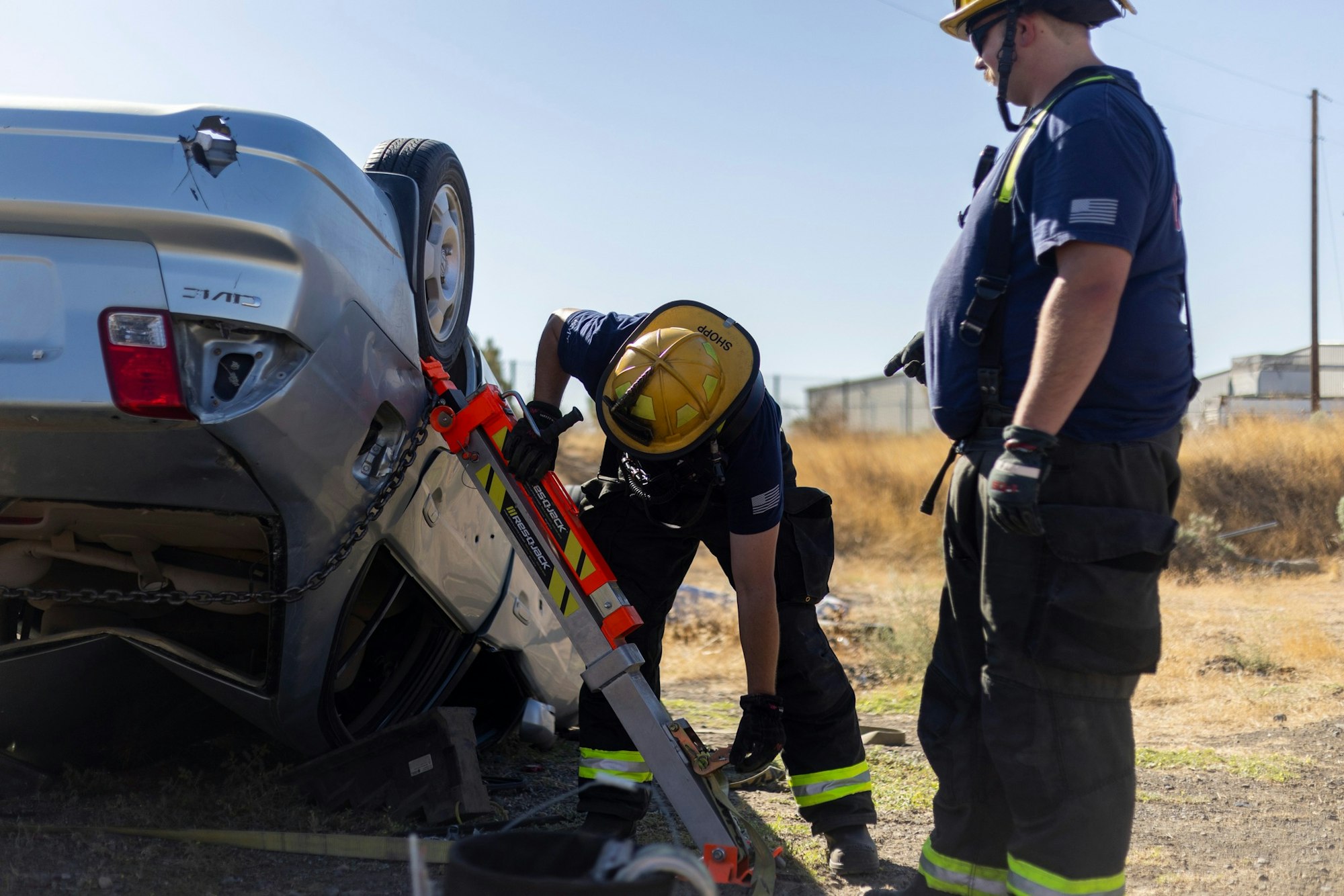 Firefighters stabilize an overturned car using equipment on a sunny day.