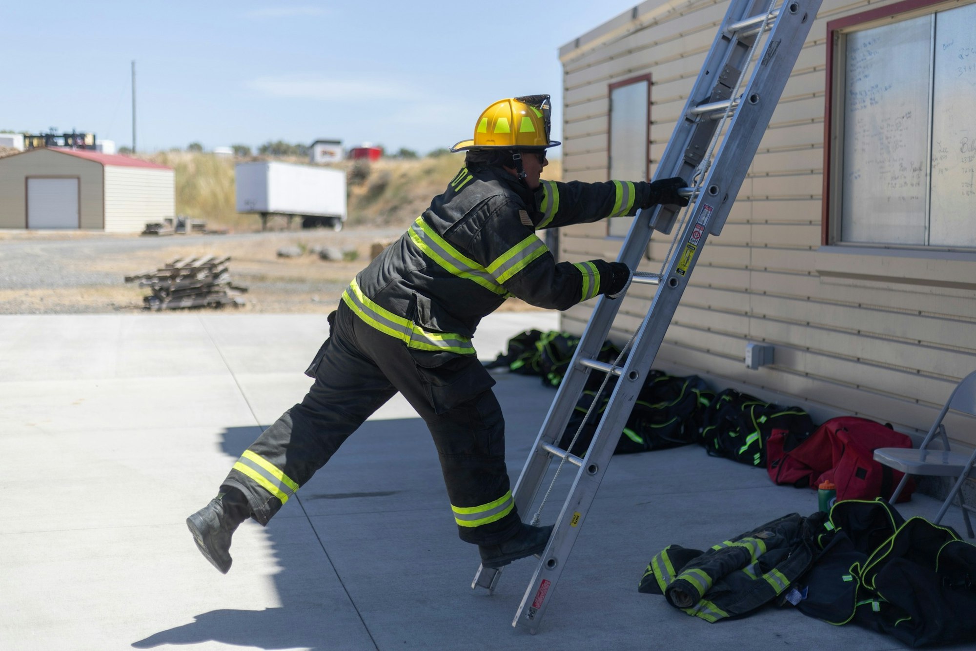 Firefighter climbing a ladder leaning against a building, wearing protective gear and a yellow helmet.