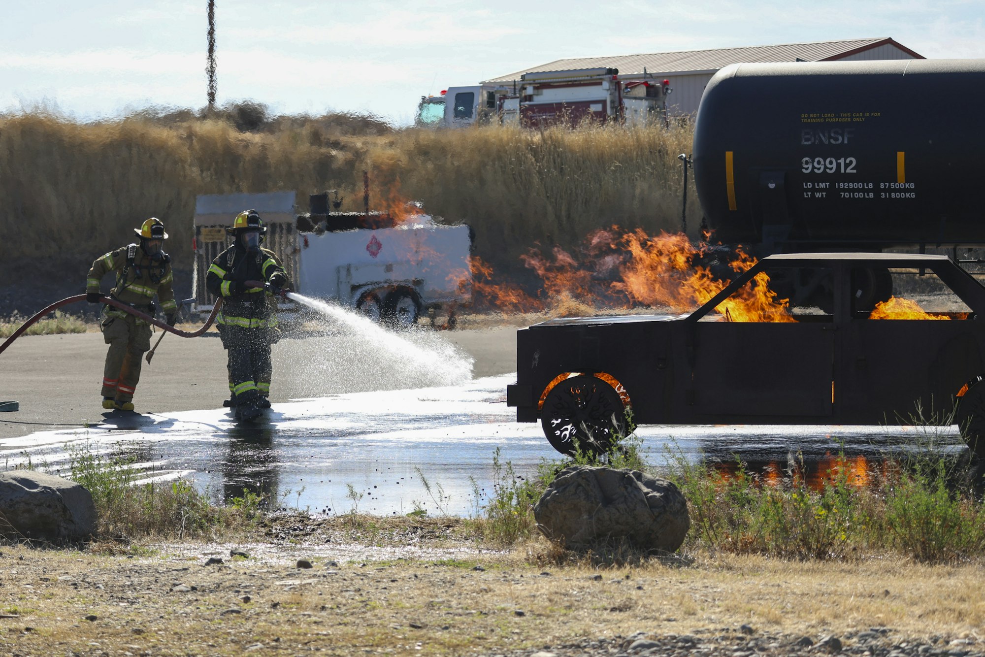 Firefighters extinguishing a training car fire with hoses, flames visible.