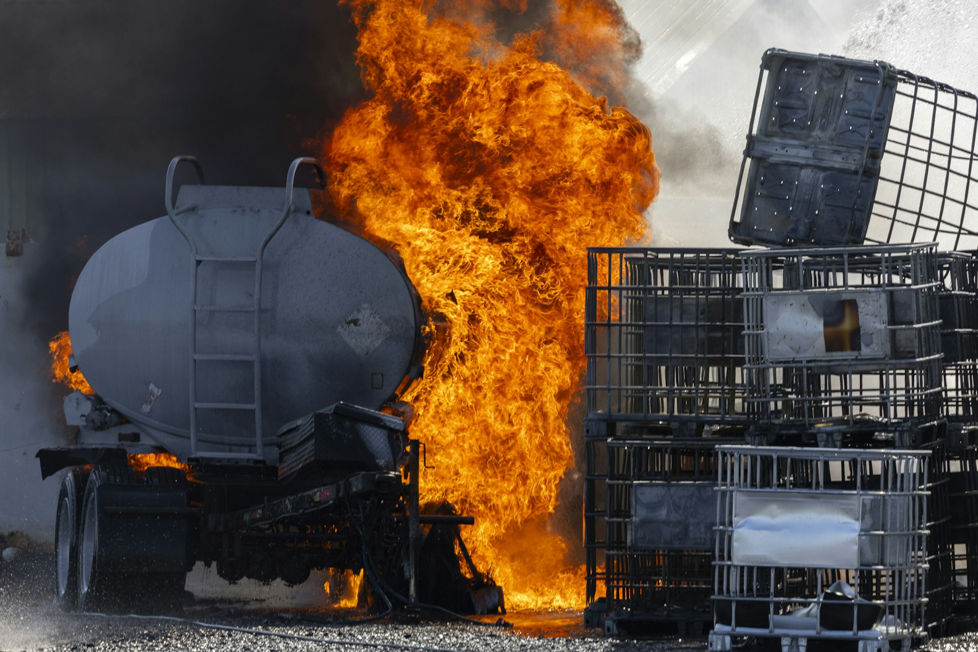 A large fire engulfs a tanker truck and nearby crates, producing thick smoke.