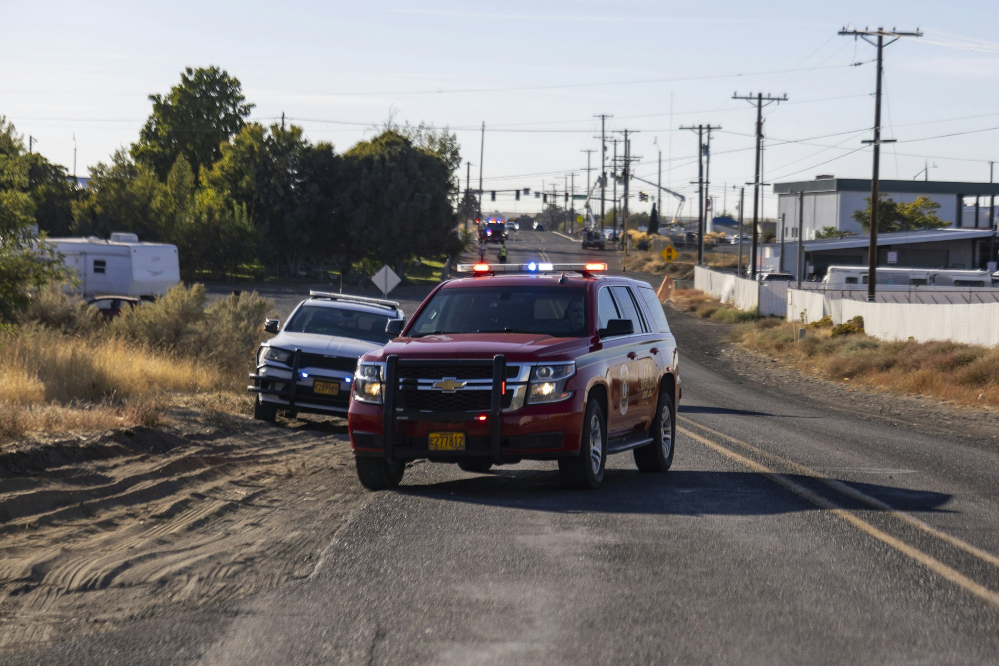Emergency vehicles with flashing lights are parked on a rural road, with a police car and a red SUV in the foreground.