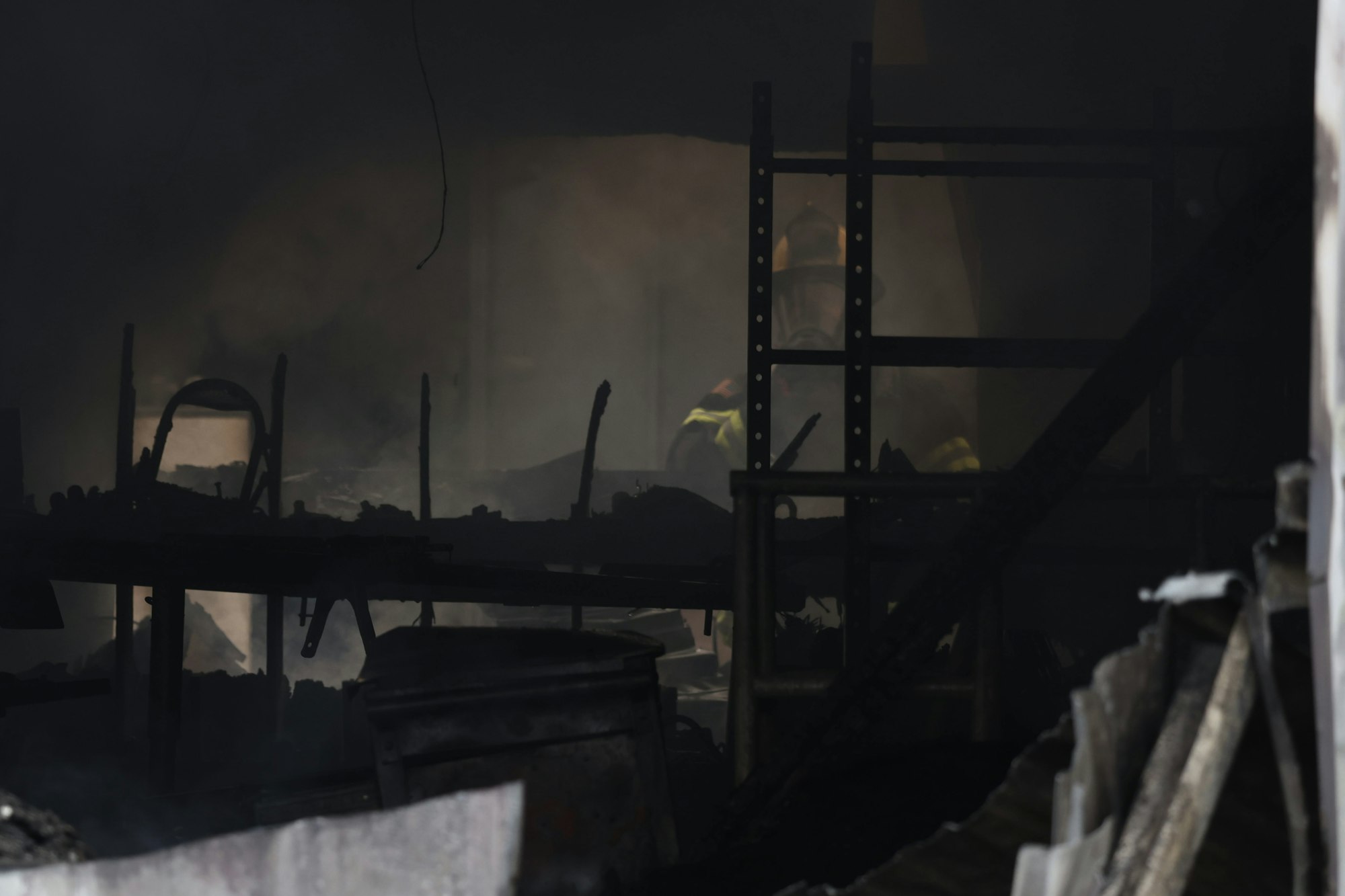 A firefighter in gear stands amidst smoke and debris inside a dark, burnt structure.