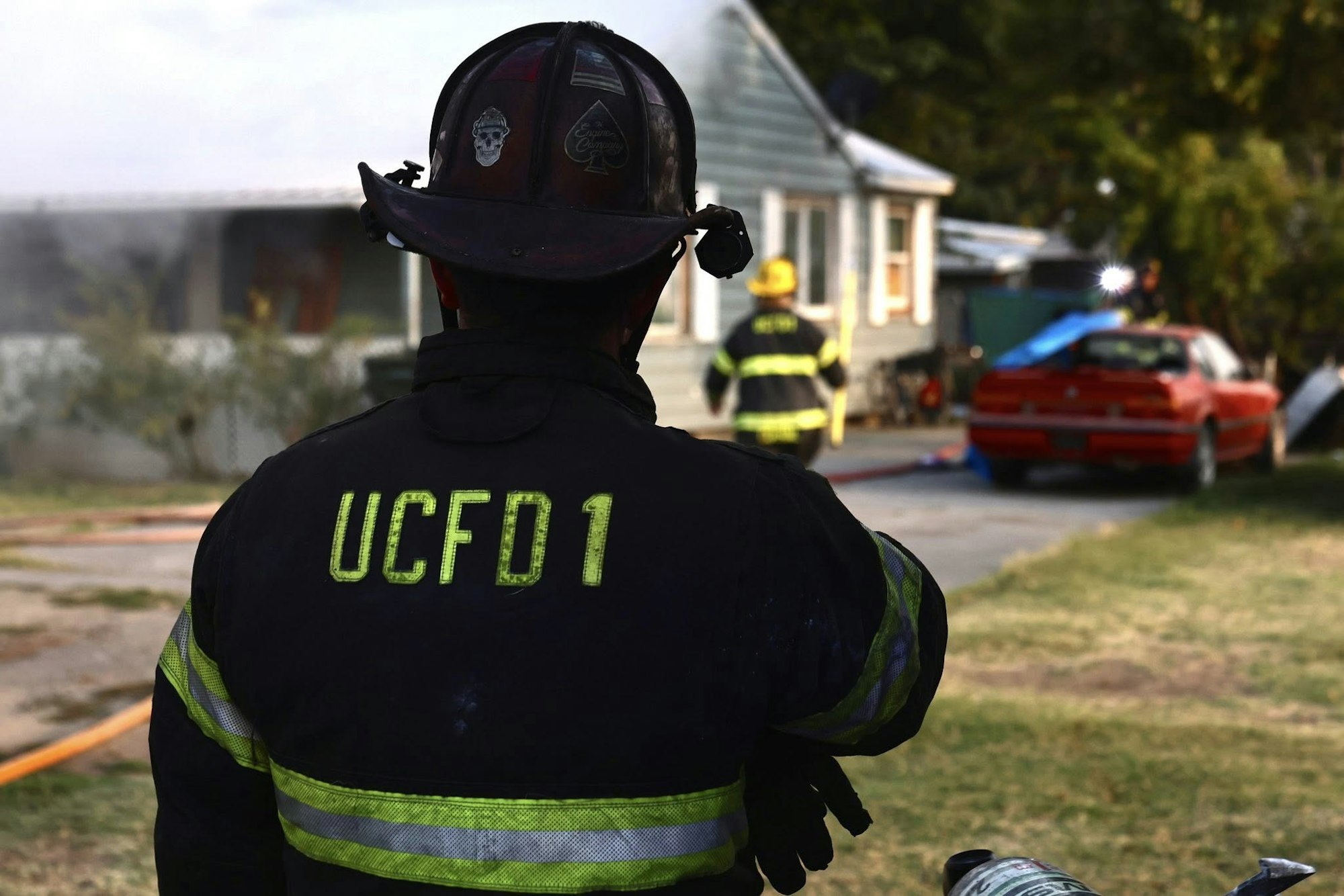 Firefighters in gear near a house with smoke; a red car nearby.
