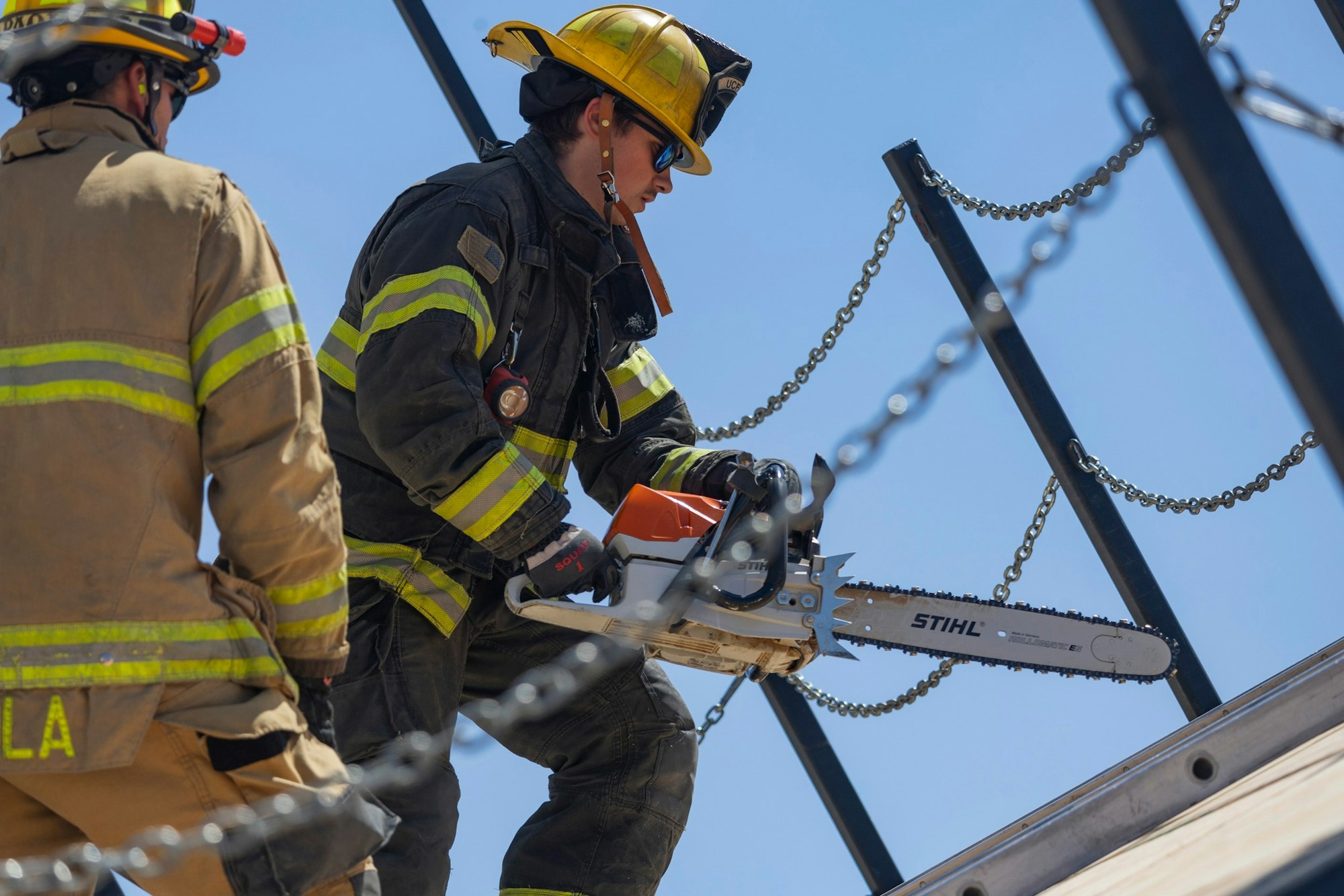 Two firefighters, one using a chainsaw, are working on a roof amidst chains and poles, wearing protective gear.