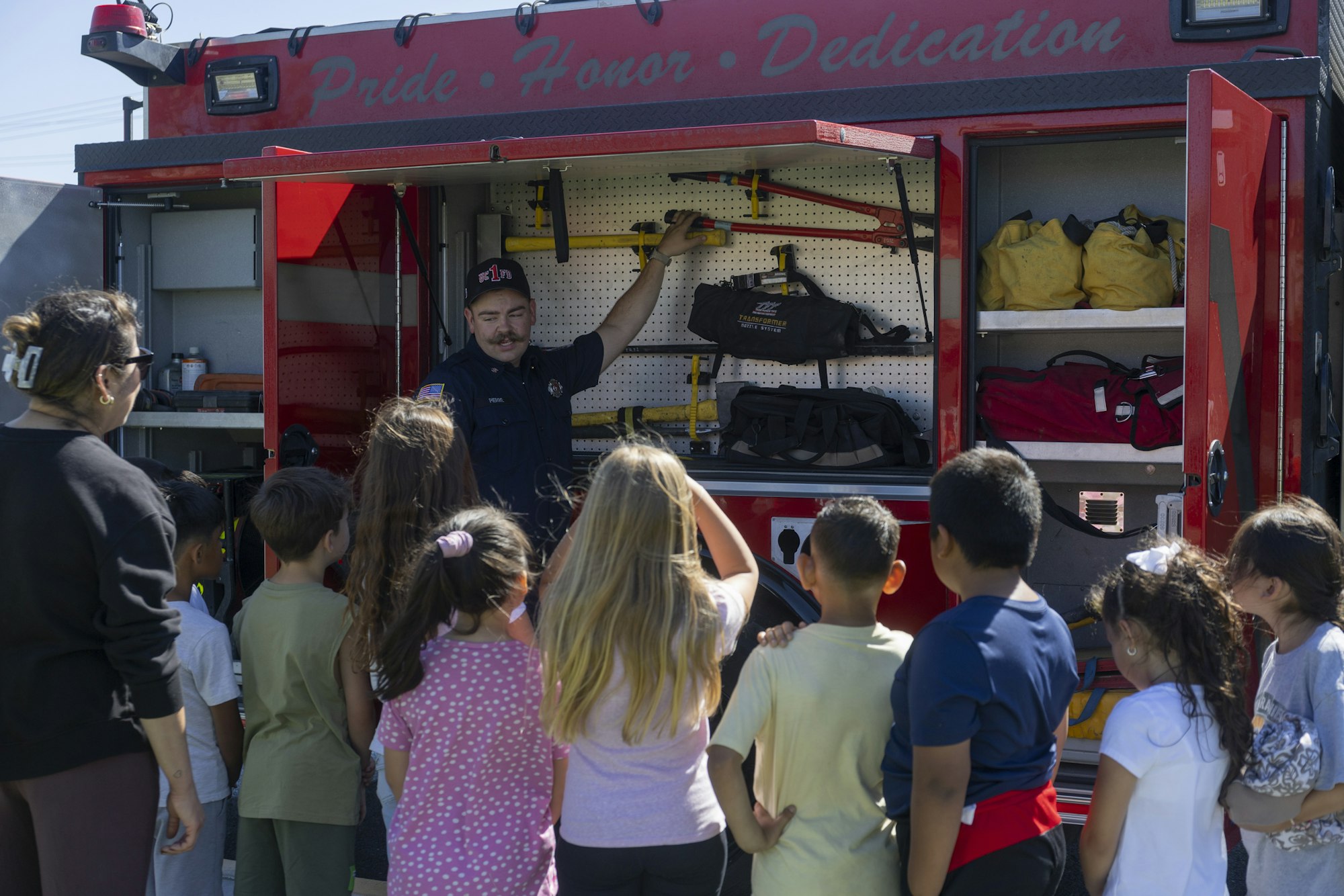 A firefighter shows tools to children standing in front of a red fire truck with open compartments.