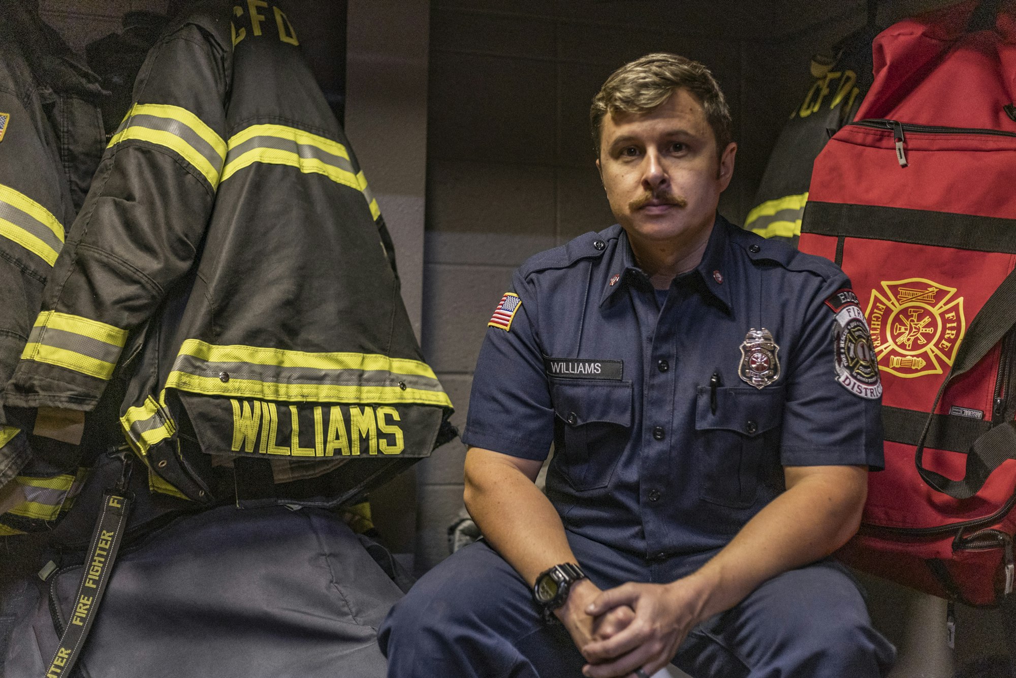 A firefighter in uniform sits by gear labeled "Williams" and a red bag with a fire emblem.