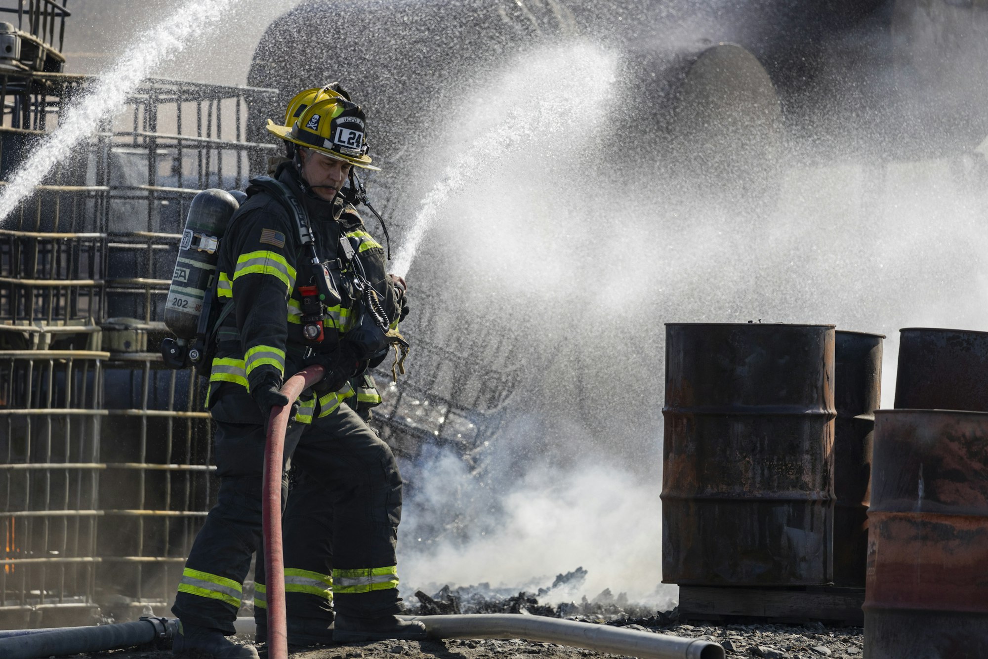 Firefighter spraying water with a hose amidst smoke and barrels.