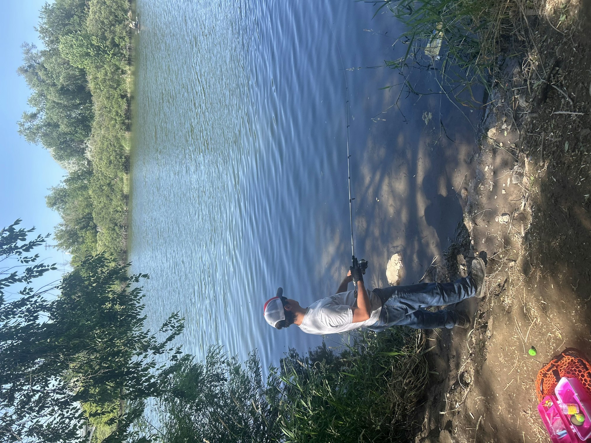 Person fishing by a lakeside, surrounded by greenery; equipment and a pink tackle box nearby.