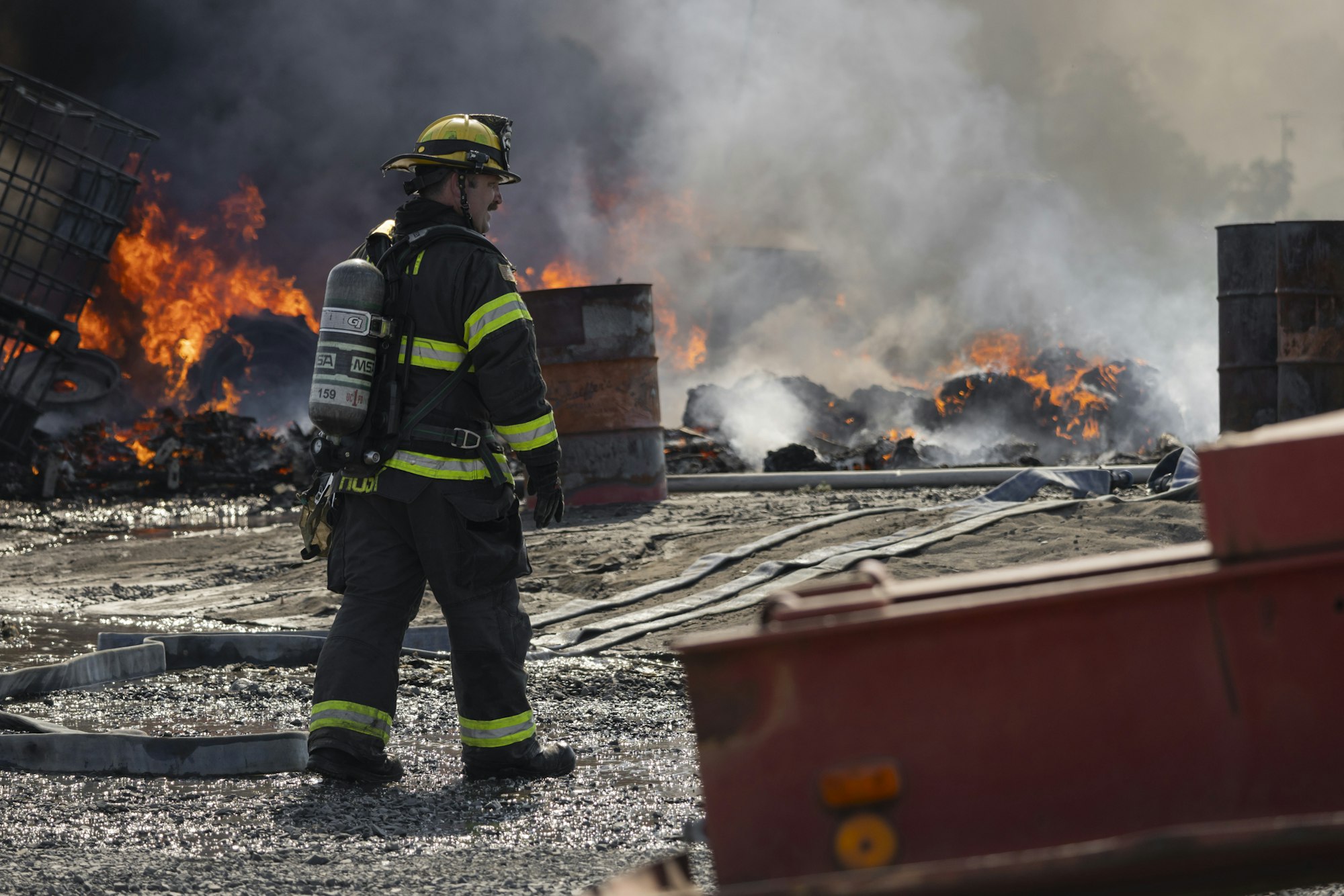 Firefighter in protective gear stands amidst a smoky, fiery scene with barrels and debris burning.