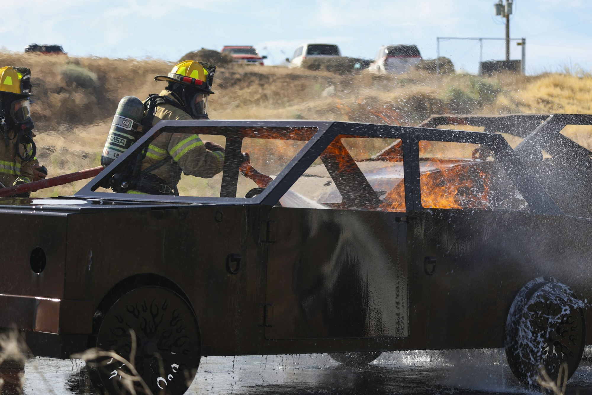 Firefighters extinguishing flames in a car using a hose, with smoke and water spray visible.