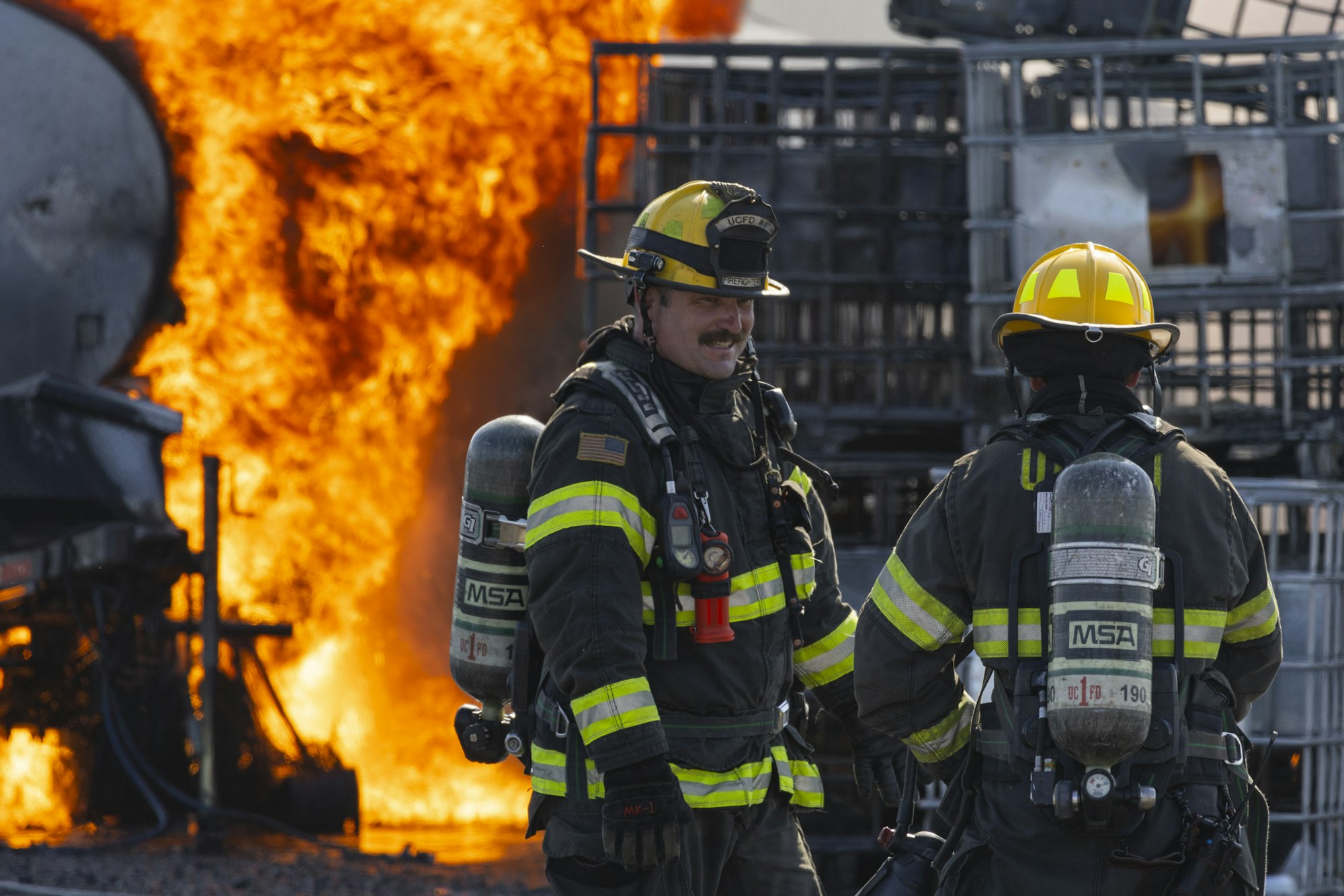 Two firefighters in gear, with helmets and oxygen tanks, stand near a large fire.