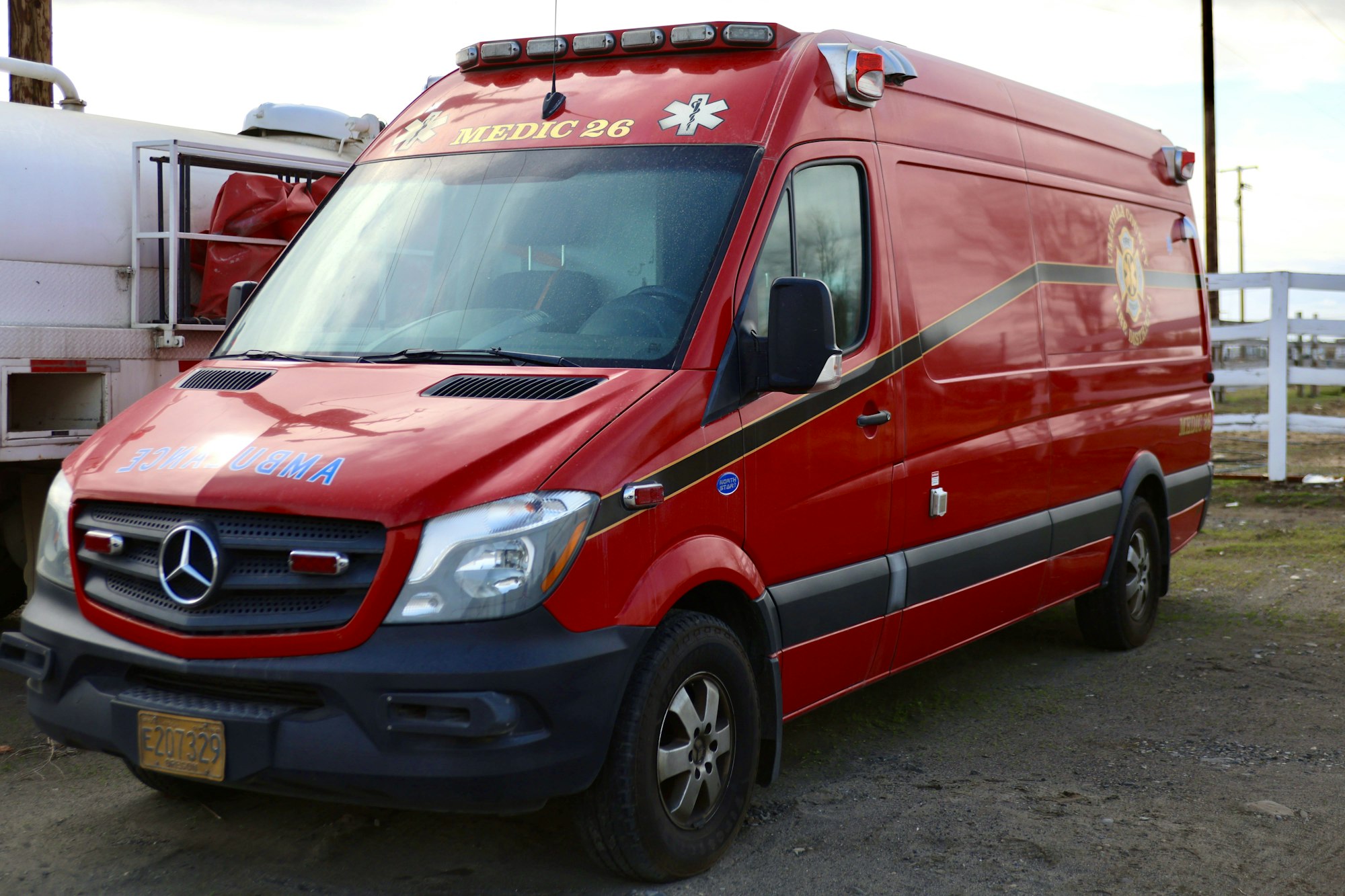 Red ambulance labeled "Medic 26" parked on gravel, with a Mercedes-Benz logo on the front.